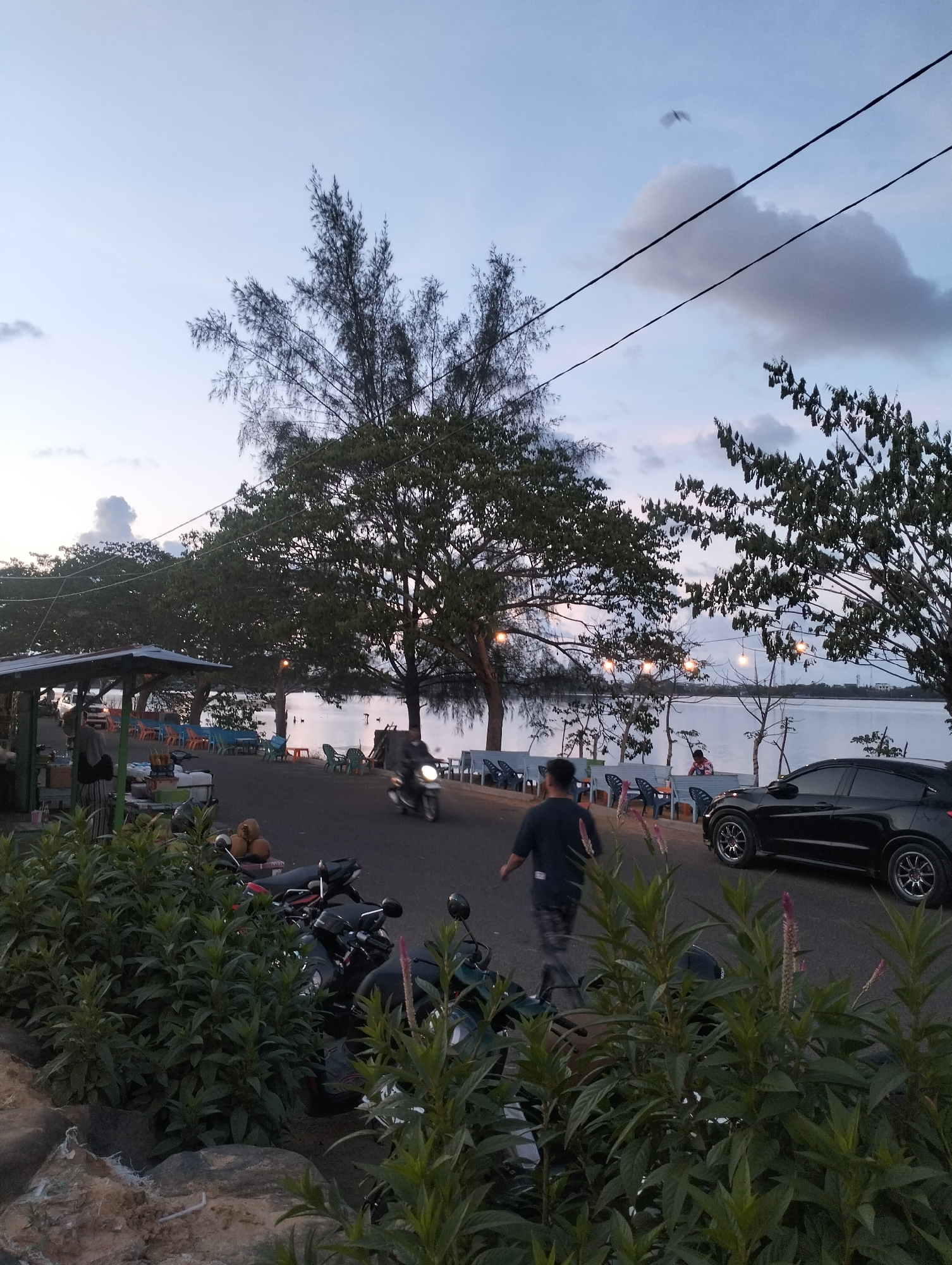 The image presents a twilight scene on a road by a body of water, potentially the sea or a large lake, in Lhokseumawe, Indonesia. A road curves through the scene, lined with foliage in the foreground. A dark-colored car is parked on the road's edge, while a person in dark clothing walks along the road. A scooter drives past. Beyond the road, chairs and tables are arranged along the waterfront. In the distance, lights are strung between trees near the water. The sky is dusky with clouds. Power lines run across the sky. There are trees lining the road. A man is sitting on the edge. Several motorcycles are parked behind the road.