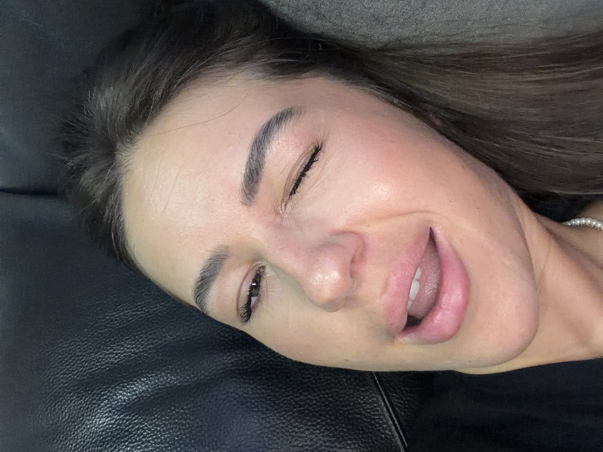 A close-up, slightly angled, eye-level shot features a woman lying down, propped up by what appears to be a black leather surface on the left and a gray textured surface on the right. The woman is winking and sticking her tongue out slightly, with her mouth open and teeth visible. Her makeup is applied, including noticeable mascara and eyeliner on her visible eye, and a pinkish hue on her lips and cheeks. Her dark hair frames her face. The lighting is soft, highlighting the contours of her face and creating a subtle sheen on her skin. The environment is dark, with only the woman's face and parts of her body and the supporting surfaces illuminated. There are no discernible landmarks or identifying features of Novi Iskar, Bulgaria in the image.