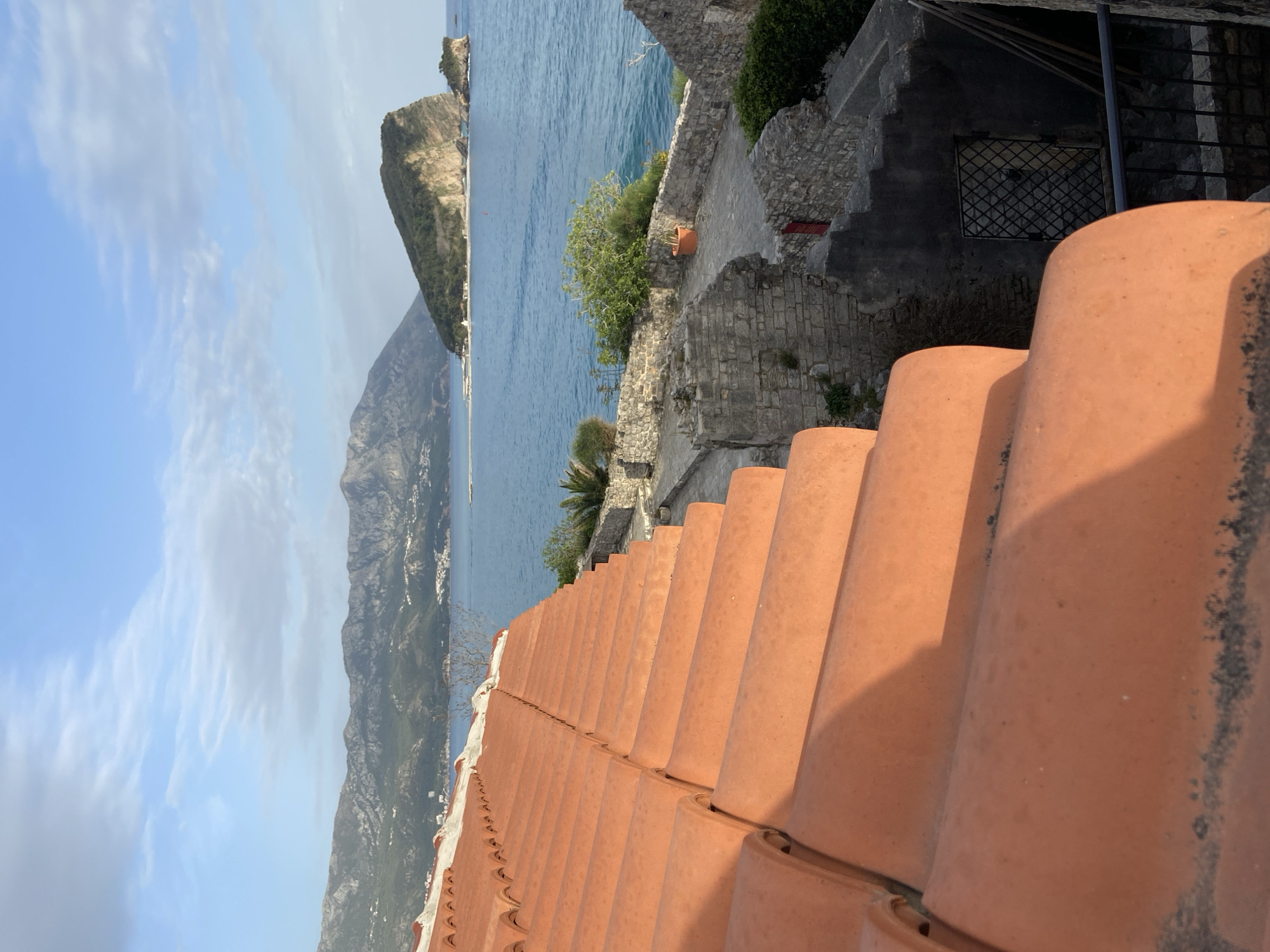 A slightly angled eye-level shot captures a portion of the red terracotta tile roof that leads to a stone staircase of an old building. To the left, the Adriatic Sea is visible, with mountains in the background. In the distance, a cliff on the sea is noted, with the sky showing light clouds. The location is near Budva, Montenegro.