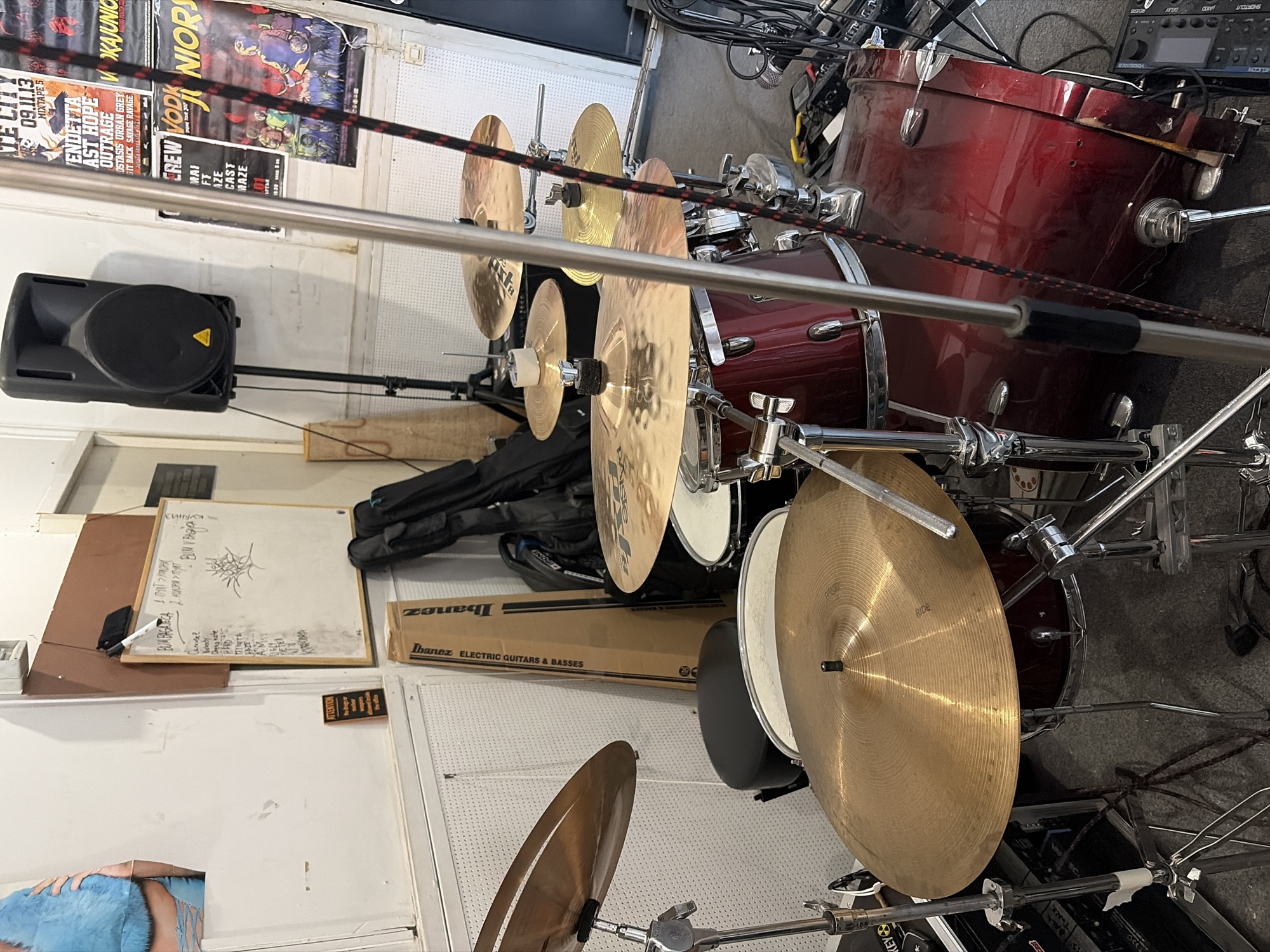 A rehearsal space or small venue in Sofia, Bulgaria, is depicted, dominated by a drum kit. The drum kit is predominantly red and appears to be in use or recently used, with several cymbals visible. The background shows a whiteboard with writing, a large speaker on a stand, and various posters on the wall, some of which are music-related. There are also instrument cases and other musical equipment scattered around. The lighting suggests an indoor setting, possibly during daytime with artificial lighting. No people are directly visible in the frame. The overall atmosphere suggests a functional, lived-in space dedicated to music practice or performance.