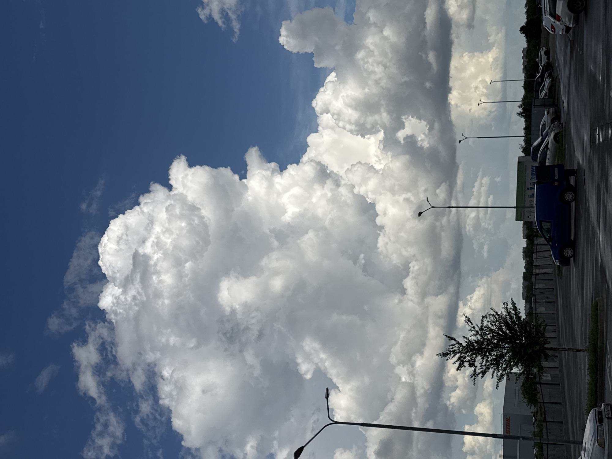 The image shows a large cumulus cloud in a blue sky, juxtaposed with a parking lot in Krasnodar, Russia. The parking lot contains several parked cars, light poles, and a single tree. The sky is partly cloudy.