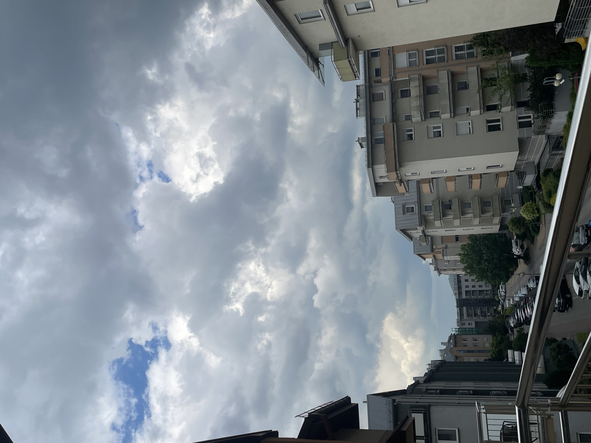 The image shows a view from a balcony in an urban environment. The sky is predominantly cloudy, with patches of blue visible through the gaps. Tall buildings line the streets, with rows of windows and balconies visible. A street with parked cars runs between the buildings. The location is in an unknown city.