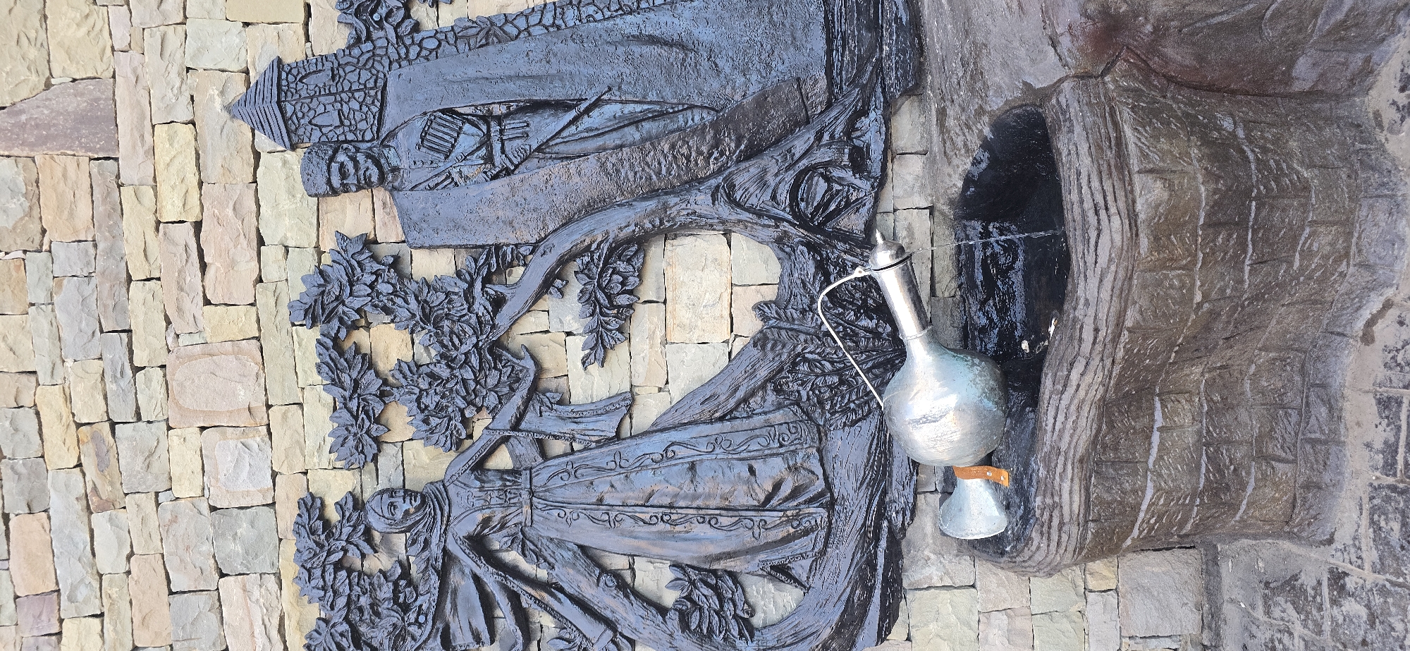 The image depicts a relief sculpture and drinking fountain integrated into a stone wall. The wall is composed of light-colored rectangular stones of varying sizes. The sculpture, rendered in a dark material resembling bronze or dark stone, features a depiction of a woman in traditional dress standing beneath a tree, with a building or castle motif above her. Water flows from a pipe into a metallic pitcher, which is supported by a separate metallic cup or base, creating a functional drinking fountain. The fountain is enclosed by a structure made of a darker, grey-toned stone. The location is identified as Grozny, Russia. There are no visible people in the image.