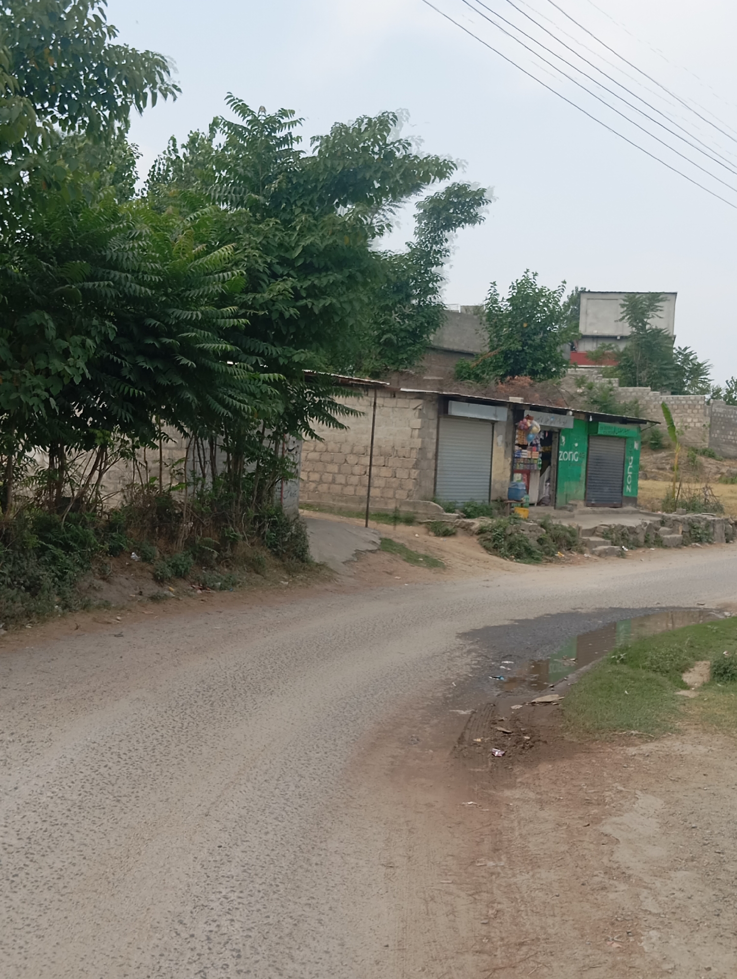 A dirt road curves through a semi-arid landscape. To the right of the road, a small shop with a green facade and roller shutters displays various goods. Two shuttered windows and an open space revealing products can be seen on the shop. The shop is built against a stone wall. Further back, buildings and trees are visible. The sky is overcast. The scene appears to be in Lassan, Pakistan.