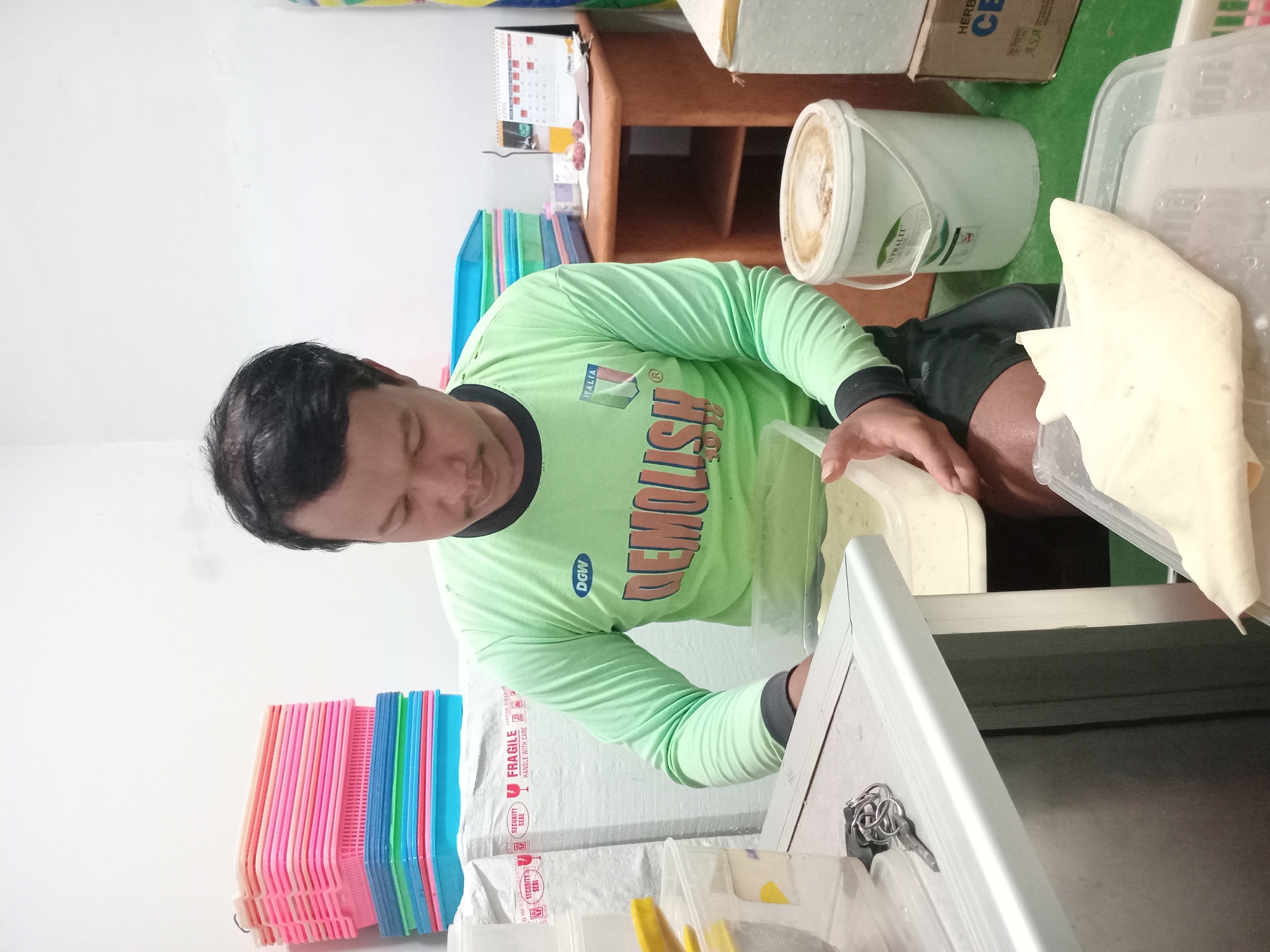 In Puncu, Indonesia, a man wearing a light green long-sleeved shirt with the text "DEMOLISH 2022" and the Italian flag logo, is seated and appears to be preparing food or a similar item. He is holding a transparent plastic container with a light-colored substance inside. In front of him is a silver-colored table with a key and keychain resting on it. Behind the man, a stack of colorful plastic baskets is visible. To his right, a wooden table holds a calendar, a glass of orange liquid, and some small red objects, possibly onions or garlic. A white bucket with a label that reads "SUPRALIT" is also present near the table. The environment seems to be an indoor workspace, possibly a kitchen or storage area, with white walls and a green floor.