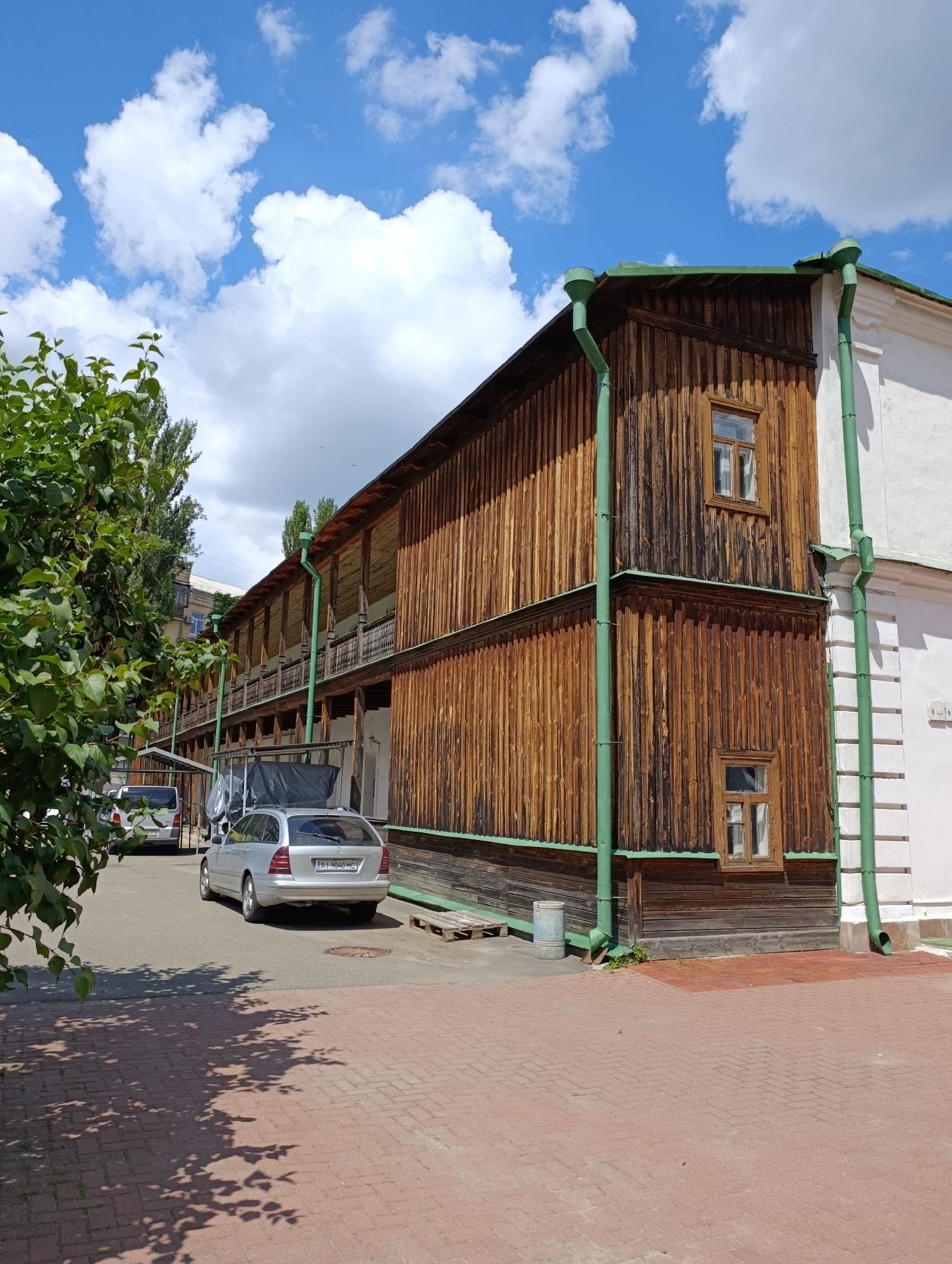 This media file depicts an outdoor scene in Kyiv, Ukraine, on a sunny day with scattered white clouds. The primary subject is a long, two-story wooden building with an open-air balcony on the upper level, supported by numerous wooden pillars. The building's facade is constructed from vertical wooden planks, and it features several small, rectangular windows with wooden frames. Green gutters and downspouts run along the exterior of the building.
To the left of the wooden structure, a section of a white building is visible, with decorative white molding along its edge. In the foreground, a brick-paved area is shaded by the leaves and branches of a large green tree situated on the left side of the frame.
In the background, parked on an asphalt surface, are two cars: a silver station wagon in the foreground and a silver minivan behind it. The station wagon has a license plate with the text "AI 9040 MC". There are also some wooden pallets and a metal trash can visible near the building. The overall impression is a quiet, daytime scene in a developed urban or semi-urban area, possibly a historical district or a residential courtyard. The bright sunlight and blue sky suggest a pleasant, warm day. No people are visible in the frame.