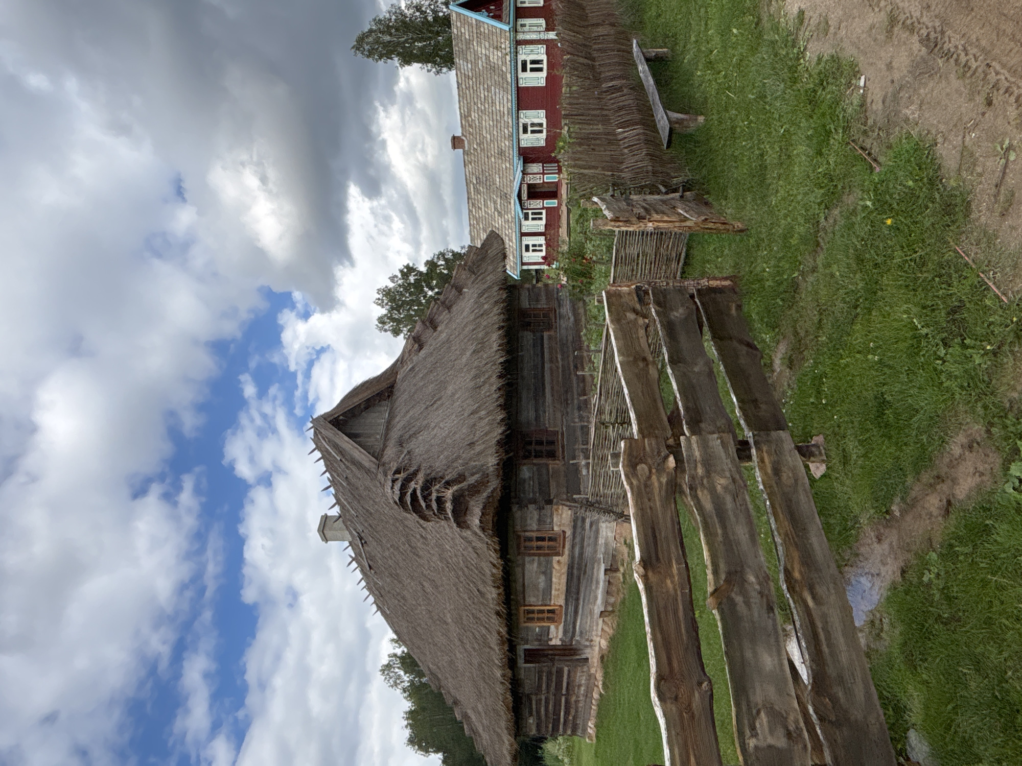 This media file shows a traditional, rustic village scene in Sielachowskie, Poland. The primary subjects are two old wooden houses. The house in the foreground has a prominent thatched roof that appears somewhat worn and damaged in places. It features small wooden windows. To the left and background is a second, larger house painted red with white trim around its numerous windows, and a grey, tiled roof. Both structures exhibit traditional architectural styles.
The setting is outdoors, with lush green grass surrounding the houses and a dirt path winding through the scene. A rough-hewn wooden fence runs along the foreground, adding to the rustic feel. The sky is partly cloudy with dramatic clouds casting shadows, suggesting a dynamic weather pattern, possibly late afternoon. There is no visible text in the image. No people or specific activities are depicted, creating a still, historical ambiance. The overall impression is one of a preserved historical village, evoking a sense of time and place.