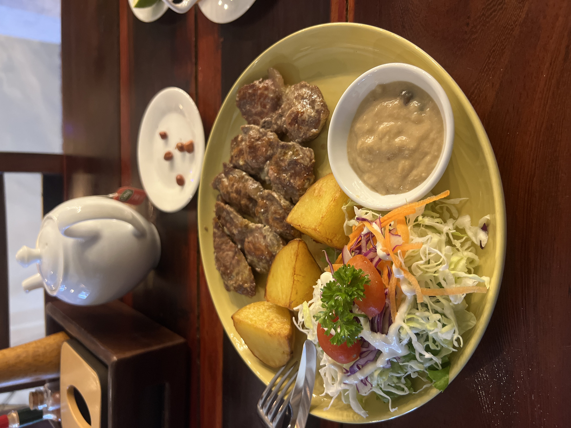 A close-up, overhead shot captures a plate of food set on a dark wooden table. The plate, a light yellow, holds a meal consisting of several pieces of grilled meat, thick-cut french fries, and a side salad. The meat appears to be tender and is coated in a creamy sauce. The fries are golden brown and have a hearty, rustic cut. The salad is a mix of shredded cabbage, carrots, and sliced tomatoes, garnished with parsley and drizzled with a white dressing. A small white bowl containing a creamy dipping sauce is positioned to the upper right of the plate. To the left of the main plate, a small white dish contains a few scattered peanuts. Further to the left, a white ceramic object, possibly a sugar bowl or creamer, sits on a dark wooden surface. The overall scene suggests a meal being served at a restaurant. The lighting is warm, implying an indoor setting, possibly in the evening or on a cloudy day, given the lack of strong sunlight. The background is dominated by dark wood, adding to the restaurant ambiance. No visible text is present in the image.