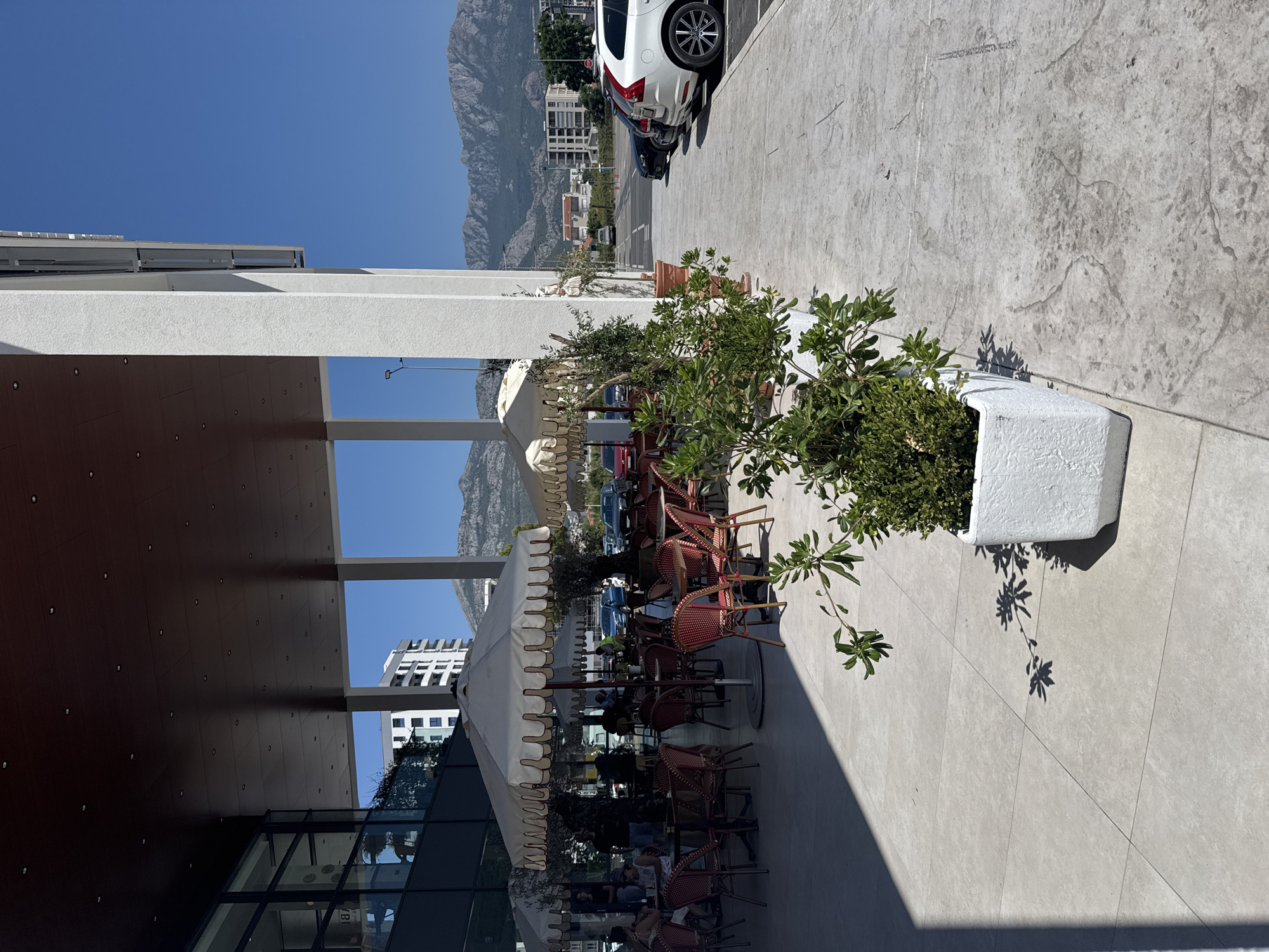 A bright, sunny day is captured from a low-angle perspective, showing a modern building's exterior with an outdoor seating area and a parking lot. The foreground is dominated by a paved walkway and planters with greenery. In the middle ground, an outdoor cafe with white umbrellas and red-and-white checkered chairs is set up on a tiled patio. People are visible sitting at tables. Beyond the cafe, a parking lot stretches out, with several cars parked, including a white Volvo SUV on the right. The background features buildings and a range of mountains under a clear blue sky. The scene appears to be in Bjeliši, Montenegro.