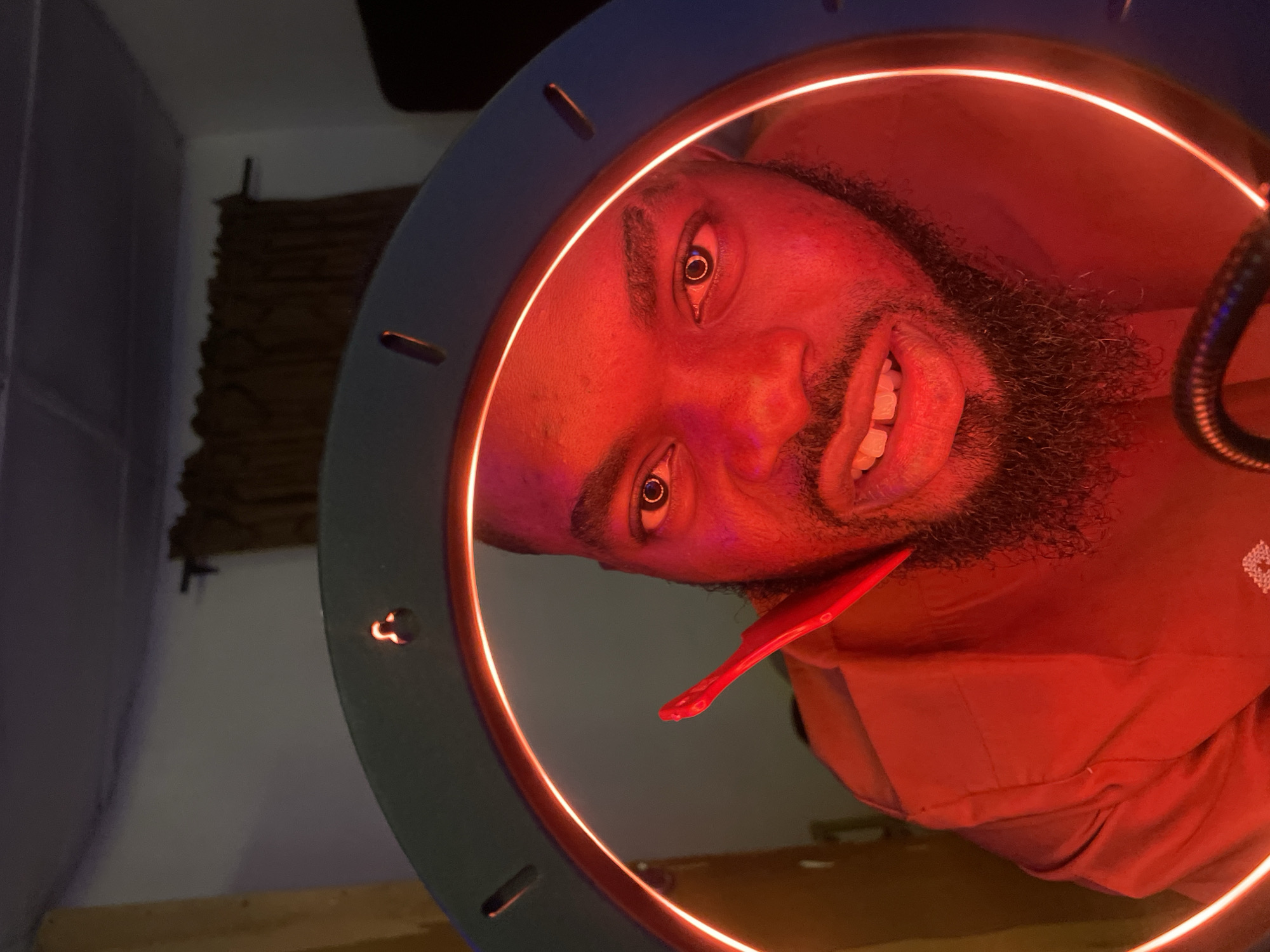 A close-up, angled portrait features a smiling person, appearing to be a male with dark skin and a beard, positioned within the bright red glow of a ring light. His head is tilted, and he looks directly at the camera with wide, expressive eyes and a broad smile, revealing his teeth. The intense red illumination bathes his face and visible upper body, creating a dramatic, vibrant hue.
The dominant feature is a large circular ring light with a dark blue or grey exterior, emitting a striking red light from its inner circumference. A small, red, spatula-like object is held near the person's chin. To the far right, a segment of a black, flexible gooseneck arm, possibly for a microphone or a small light, is visible.
The background is dimly lit and consists of a plain light-colored wall on the left and a darker, textured brown element, possibly a woven wall hanging or curtain, further back. A hint of a wooden surface can be seen at the bottom right. The overall scene suggests an indoor setting, likely a studio or a space set up for photography or video recording. There is no visible text in the image.