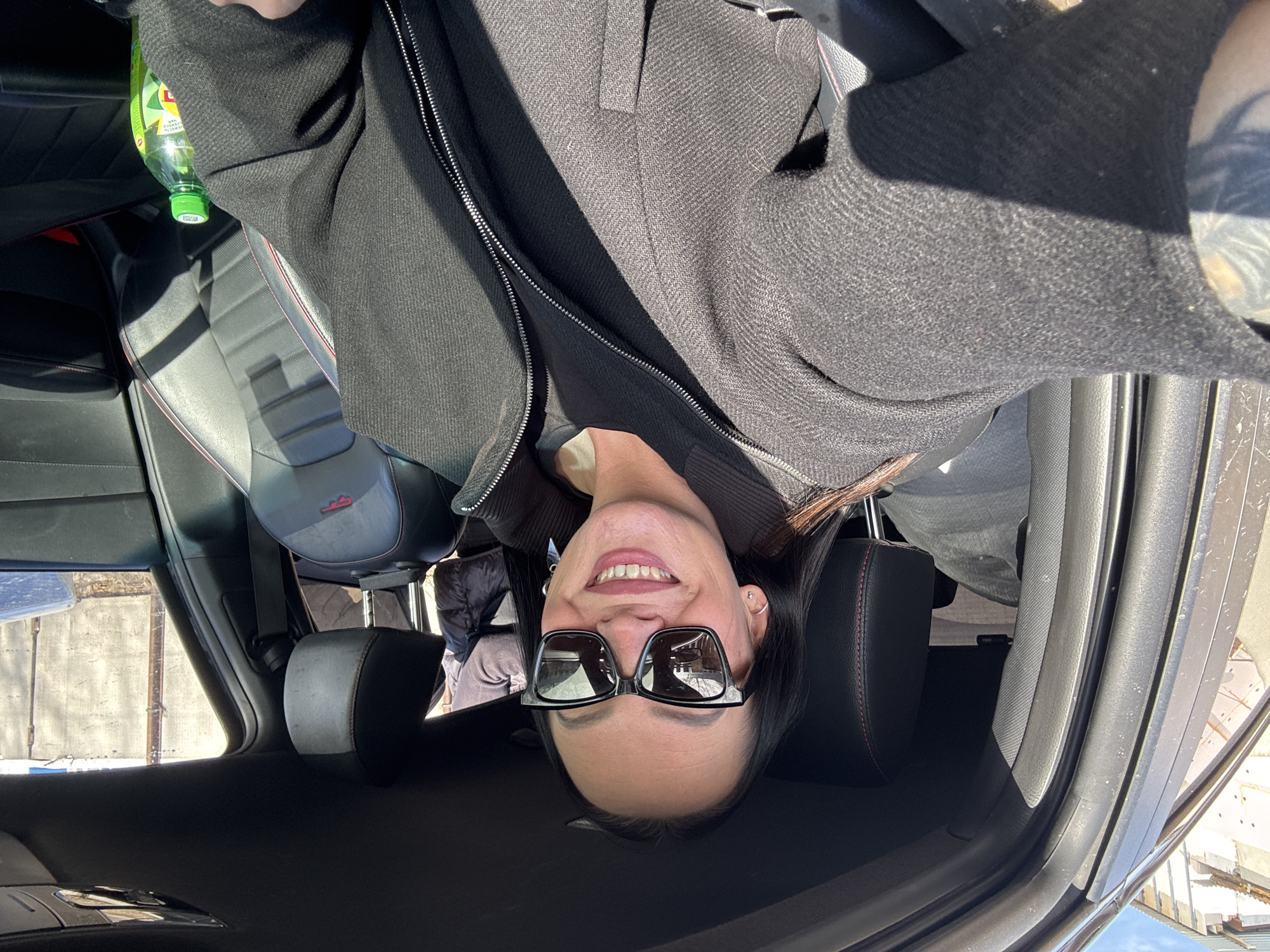 A female individual is depicted in a close-up selfie from the driver's seat of a vehicle in Chaikovsky, Russia. She has long, dark hair, wears dark, square-framed sunglasses, and is smiling with visible teeth. Her attire includes a dark grey zippered jacket over a black undershirt. A tattoo is visible on her left forearm, and a silver stud earring is present on her left earlobe. The vehicle's interior features black seats with red stitching and a visible "GT" logo on the front passenger seat. A green plastic bottle with a yellow label is located on the front passenger side. A second individual, wearing a dark puffy jacket, is partially visible in the rear passenger compartment, accompanied by light-colored items. The external environment, viewed through the car windows, shows concrete structures, a light-colored wall or fence, and a clear blue sky, indicating a sunny daytime setting.