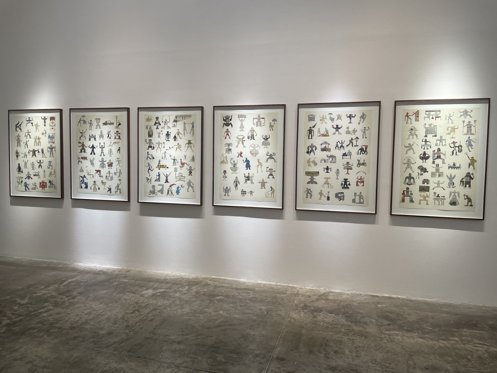 The image shows a series of six framed artworks displayed on a white wall in what appears to be an art gallery or museum. The artworks are lined up horizontally. The location is presumably in Mexico City, Mexico, though this information is not directly evident from the image itself. The artworks consist of numerous small, seemingly abstract or stylized figures and symbols arranged on a white background, each framed in a dark brown or black frame. The floor is a polished concrete surface, reflecting light. The lighting in the room is a mix of natural and artificial, with shadows cast from the artworks onto the wall. There are no people visible in the frame.