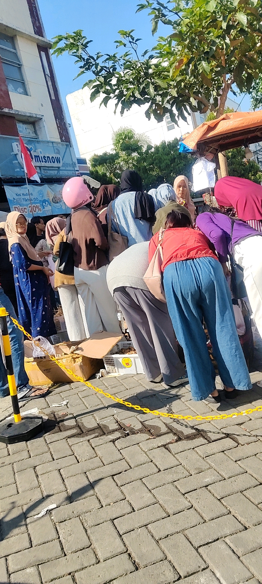 In the city of Cibatu, Indonesia, a group of people are gathered around a street-level stall. The stall appears to be selling produce, with several crates of what seem to be potatoes visible. Some individuals are leaning in, examining the goods, while others stand nearby. Among the shoppers, one person wears a bright pink helmet. The scene is set against a backdrop of buildings and trees, under a clear blue sky. A red and white flag, possibly the Indonesian flag, is visible on a building in the background. A yellow plastic chain delineates a boundary in front of the stall. The ground is paved with interlocking bricks, and shadows suggest it is daytime.