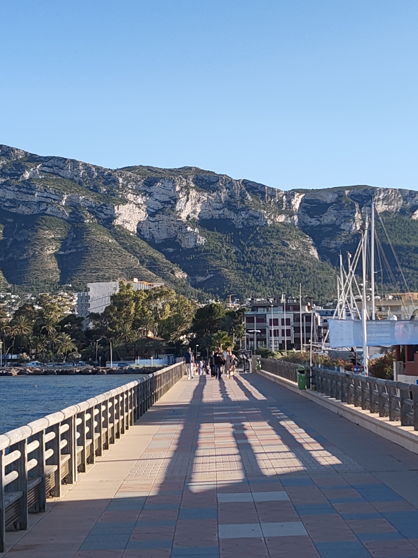 The image depicts a sunny day in Dénia, Spain, on a pier with a tiled surface leading towards the city. A group of people are walking on the pier. The pier is lined with a wooden railing on both sides. Shadows from the railing stretch across the pier's surface. In the background is a mountain range. A mix of buildings and trees are visible on the shoreline near the base of the mountains. The sky is clear and blue.