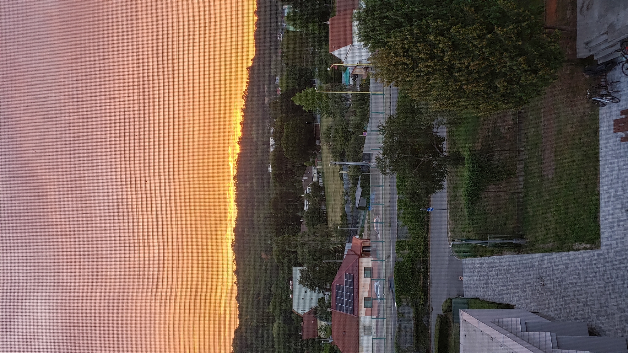 The image is a vertical, medium-shot photograph taken from a slightly elevated perspective, looking out through a window screen. The scene depicts a suburban or semi-rural landscape at sunset. The sky is a gradient of warm orange and pink hues, with the sun low on the horizon behind a silhouette of trees and a hill.
In the middle ground, there are several buildings visible. A prominent red-roofed house with solar panels on its roof is in the left-center. To its right, a white building with a red roof is partially visible. Behind these houses, a line of trees forms a dense green backdrop, with a gently sloping hill covered in more trees rising behind them. A field or open grassy area is visible between the houses and the tree line. A road runs horizontally across the middle of the image, with a few cars driving on it. Fencing and hedges line the road.
In the foreground, on the left, is a section of a modern building with a light-colored, angled facade and a paved walkway. To the right of the walkway is a grassy area. At the bottom right, near a concrete structure, there are two bicycles parked upright, one white and one black, and a red bicycle lying on its side. There are no people visible in the image. The overall impression is one of a quiet evening in a residential area. The specific location within Prešov, Slovakia, cannot be determined from the visual information alone.