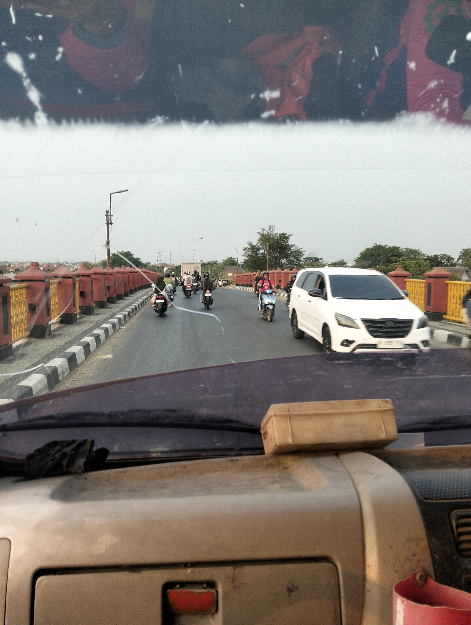 A low-angle shot looks through a cracked windshield at a two-lane road with a concrete barrier along the left side. The barrier is red and yellow with ornate posts. Several motorcycles are on the road, heading away from the camera, some with passengers. A white SUV is in the right lane, also heading away from the camera. The sky is overcast. In the foreground, the dashboard of the vehicle is visible, with a tan box on top and a black glove resting on the windshield. In the reflection of the windshield, two individuals in red shirts are visible. This scene appears to be on a bridge over a body of water, and based on the railing design and the style of the buildings visible in the background, it is likely located in Semarang, Indonesia, possibly near the Kota Lama district or along the arterial roads leading to it.