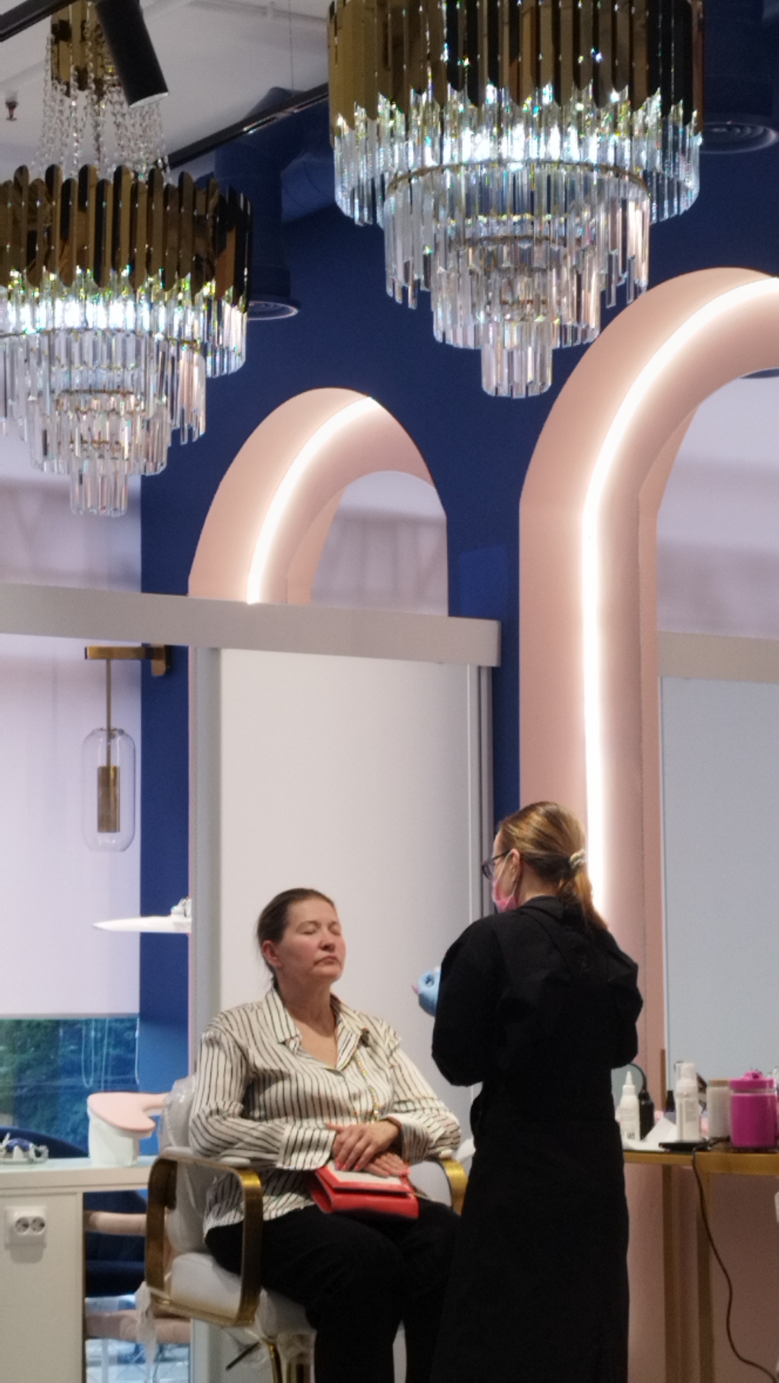 A woman sits with her eyes closed in a chair, wearing a striped shirt and black pants. She holds a red clutch in her lap. Another person stands in front of her, wearing a black outfit and a pink face mask, with their back to the camera. The room has a modern design with large arched structures and ornate chandeliers. The setting appears to be an indoor salon or spa.