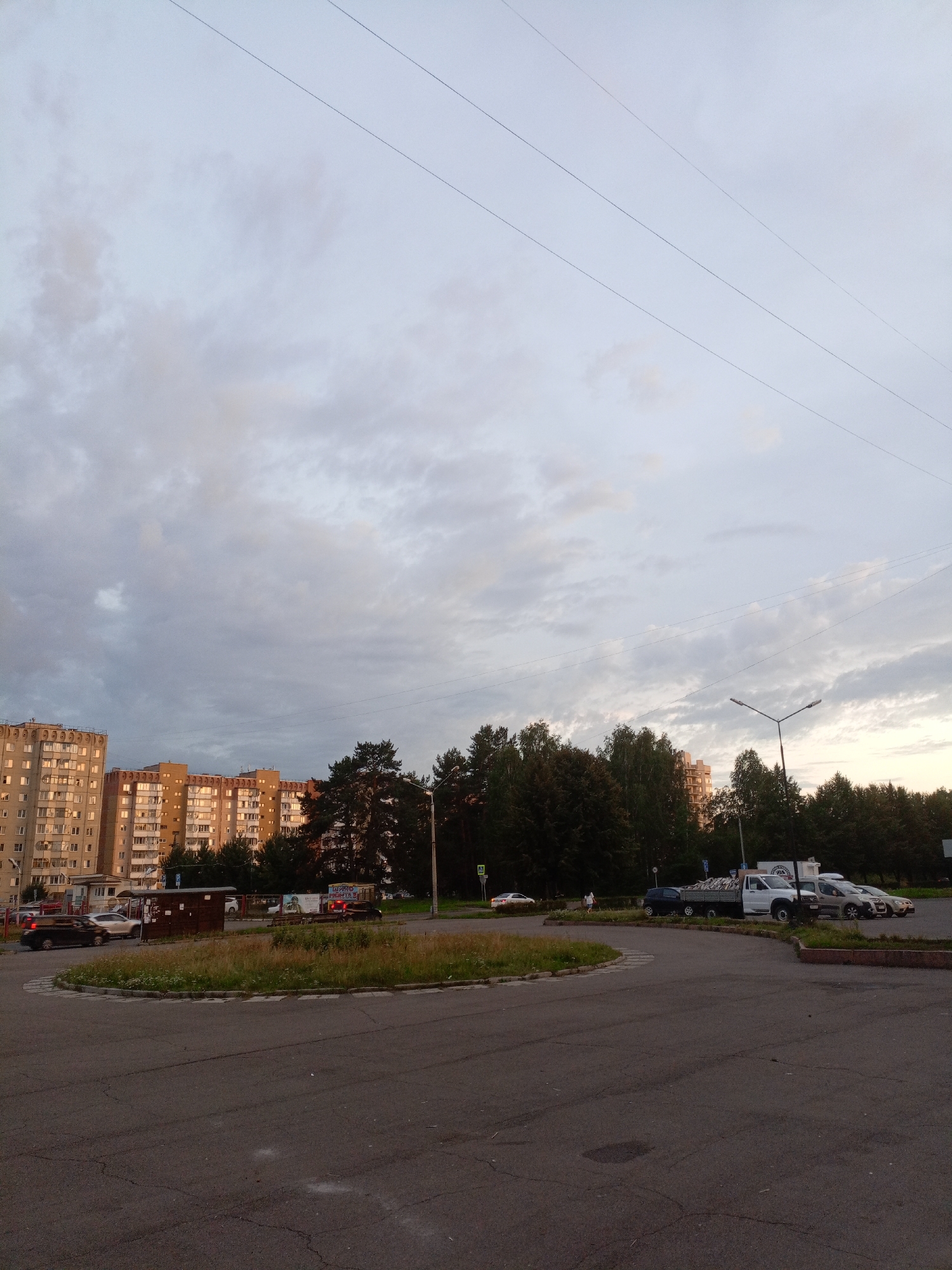 The image shows a street scene in Zheleznogorsk, Russia, during the day with overcast skies and muted lighting. In the background, two large, light-colored apartment buildings with many windows are visible. To the right of the buildings, a dense line of dark green trees forms a backdrop. A roundabout with a grassy island is in the foreground. Several cars are parked on the right side of the roundabout, including a white flatbed truck loaded with materials, a silver sedan, and a dark-colored SUV. A street light with two lamps is positioned near the trees on the right. Power lines are strung across the sky. The road surface appears to be asphalt, with some cracks and white markings. A small brown structure, possibly a kiosk or bus stop shelter, is located on the left side of the roundabout.