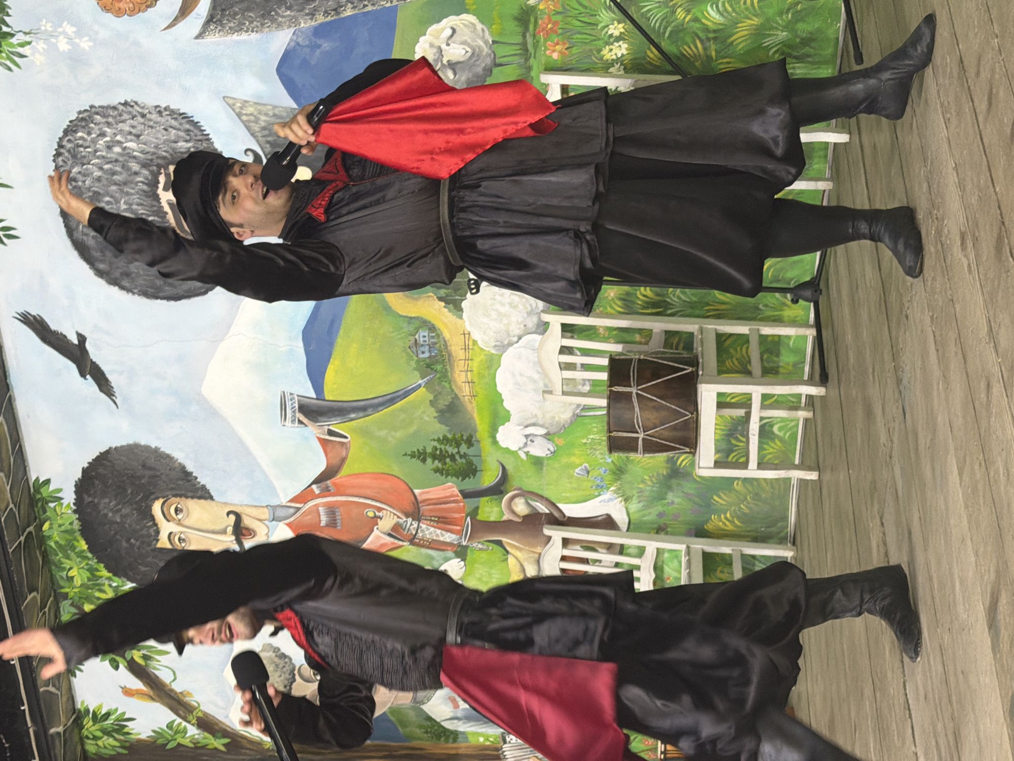 In Kveda Chkhutuneti, Georgia, two male performers in traditional black and red costumes are on a wooden stage in front of a colorful mural depicting a pastoral scene. The performer on the left, with a raised left arm, holds a microphone in his right hand and wears a black hat and a red sash. The performer on the right, also with a raised arm, holds a microphone in his left hand and a red cloth in his right, wearing a similar black hat with a feathered adornment. The mural features stylized human figures, sheep, trees, mountains, and a bird in flight. Two white chairs flank the performers, and a drum sits on a platform behind the performer on the right.