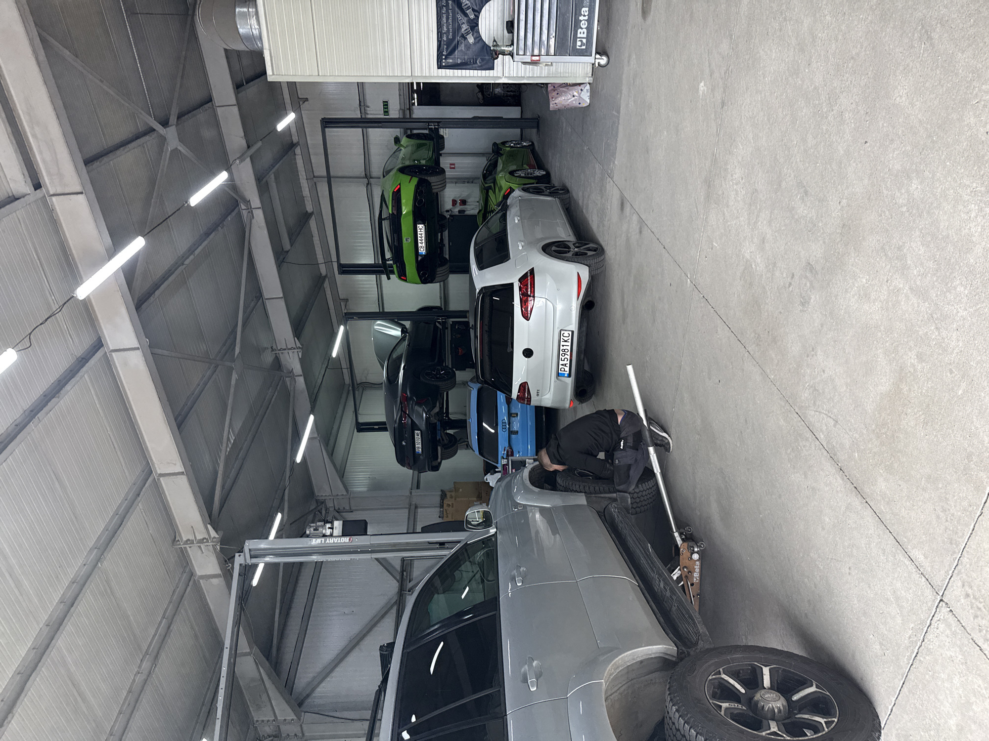 This image shows the interior of an auto repair shop with a variety of cars on lifts. There are green, white, black, and blue cars on the lifts. A man in black work clothes is working on a light grey SUV that is on the ground and jacked up. There are tires scattered around the SUV. There is a "Rotary Lift" present, along with a "Beta" toolbox. A sign in German is on a white door to the right. License plates visible include "PB 1010 ME", "CB 4444 HC", and "PA5981 KC". Based on the license plates, the location is likely Plovdiv, Bulgaria. The building has a high ceiling with metal beams and fluorescent lighting.