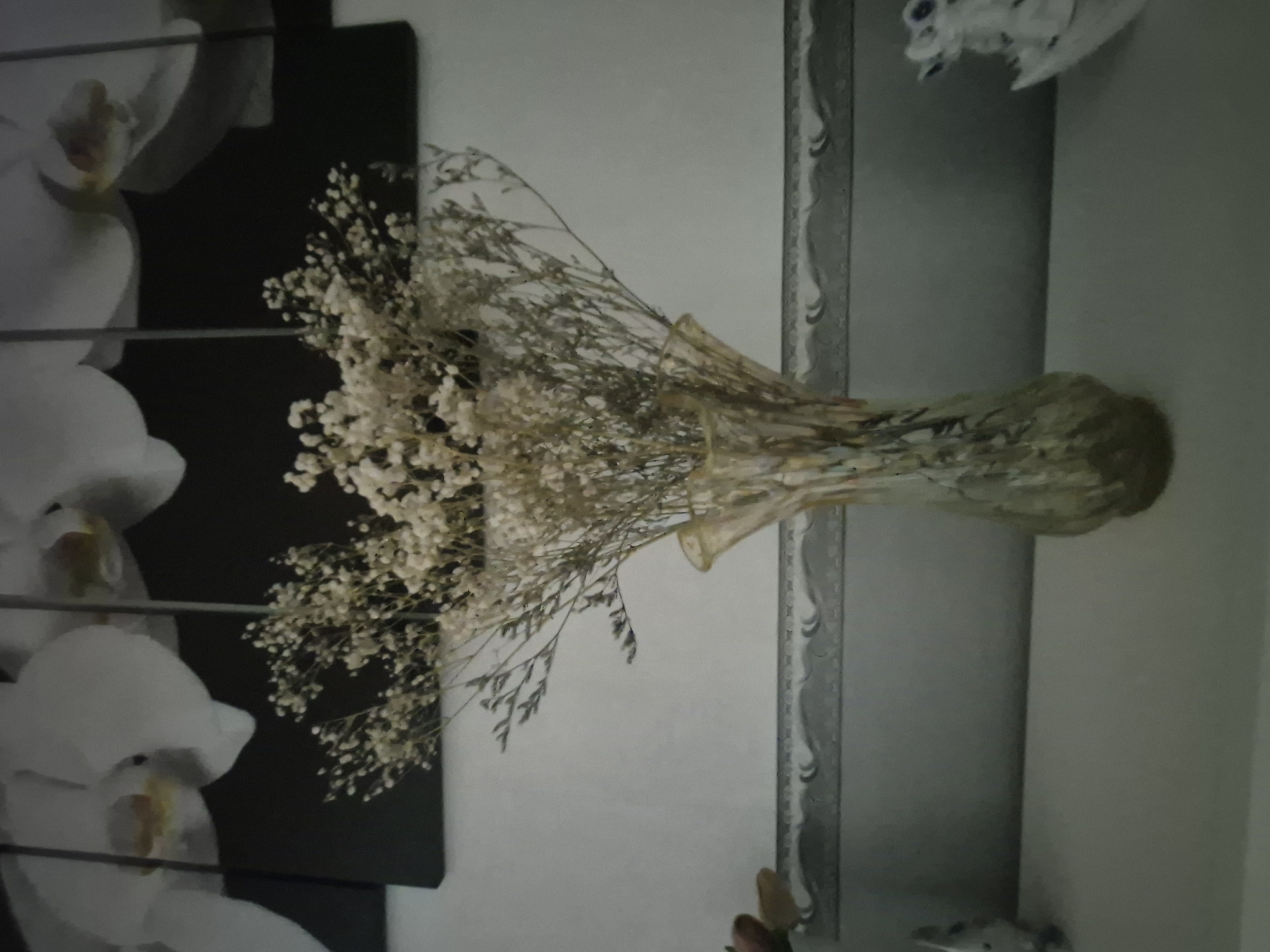 A still life photograph, taken indoors, features a vase of dried flowers placed on a flat surface. The vase is translucent and appears to be made of glass, with a decorative, possibly marbled, pattern. The dried flowers are delicate and light-colored, with many small blooms and thin stems, creating a wispy, airy arrangement. Behind the vase, a piece of artwork or a wall decoration hangs, depicting large, stylized white orchids against a dark background. Below the vase, a decorative border or trim runs horizontally across the scene. To the left and right of the vase, partially visible on the flat surface, are small decorative objects. The overall lighting is soft, and the image has a slightly hazy quality. The location is identified as Novoishimskoe, Kazakhstan.