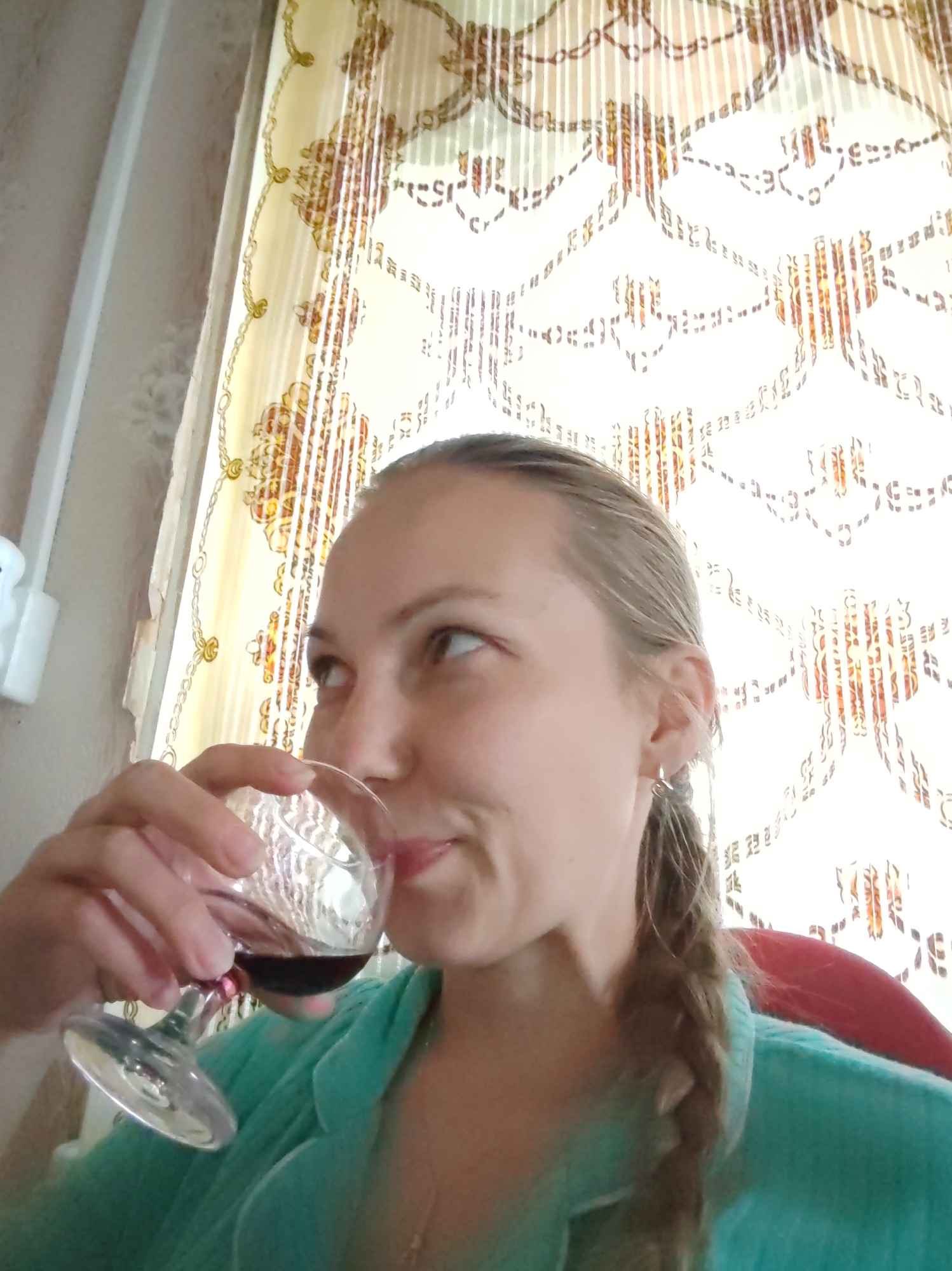 A young woman with blonde hair in a braid is holding a glass of red wine to her lips in front of a patterned curtain. She is wearing a teal colored top. The scene is likely indoors, possibly in a home in Chuhlém, Russia, given the presence of what appears to be a curtain commonly found in residential settings in the region. The lighting suggests it is daytime.