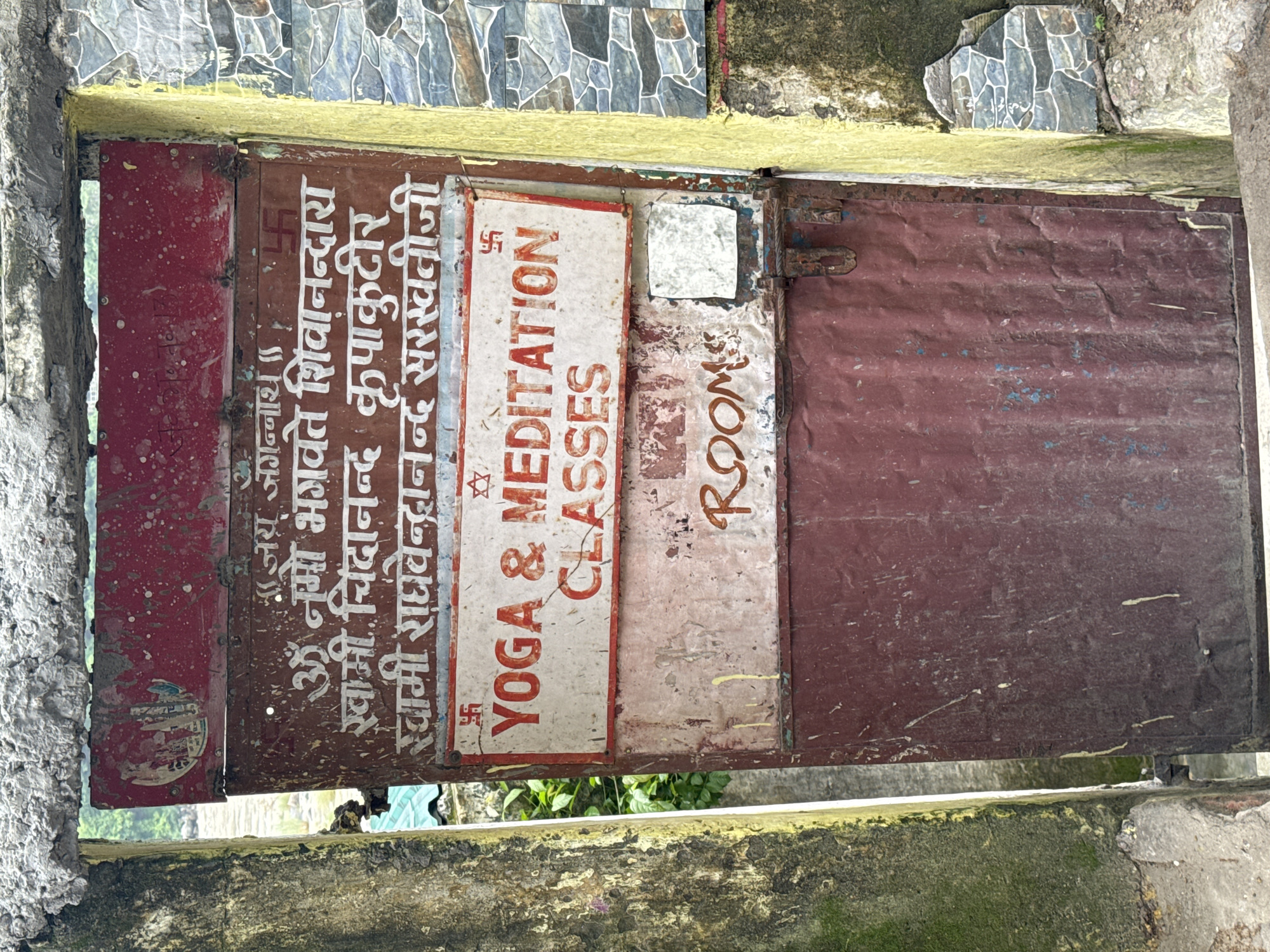 The image shows a weathered, burgundy metal door in Rishikesh, India. The door is adorned with several signs. The uppermost sign is red and appears to have some faded text and images. Below this is a brown sign with Hindi text in white lettering, preceded by a swastika symbol. The main text on this sign reads, "जय जगन्नाथ" (Jai Jagannath), followed by "ॐ नमो भगवते शिवानन्दारा" (Om Namo Bhagavate Shivanandara). The following lines are "स्वामी चिदानन्द कृपाकुटीर" (Swami Chidananda Kripa Kutir) and "स्वामी राघवेन्द्रानन्द सरस्वतीजी" (Swami Ragavendrananda Saraswatiji).
Beneath this, a white sign with a red border displays "YOGA & MEDITATION CLASSES" in red, block letters. There are also swastika and star symbols on this sign.
The lowest sign visible on the door has the word "ROOMS" in large, orange, distressed lettering. A small, reflective square is affixed to the door below this. The door itself is scratched and appears old, with a visible latch mechanism.
The door is set within a textured, concrete entryway. To the left of the door is a rough, concrete pillar covered in moss. To the right, a wall is tiled with grey and blue stones, and the surrounding concrete structure is stained with yellow and green growth. Lush green foliage is visible behind the door, suggesting an outdoor or semi-outdoor location. The overall impression is one of a rustic, spiritual establishment.
