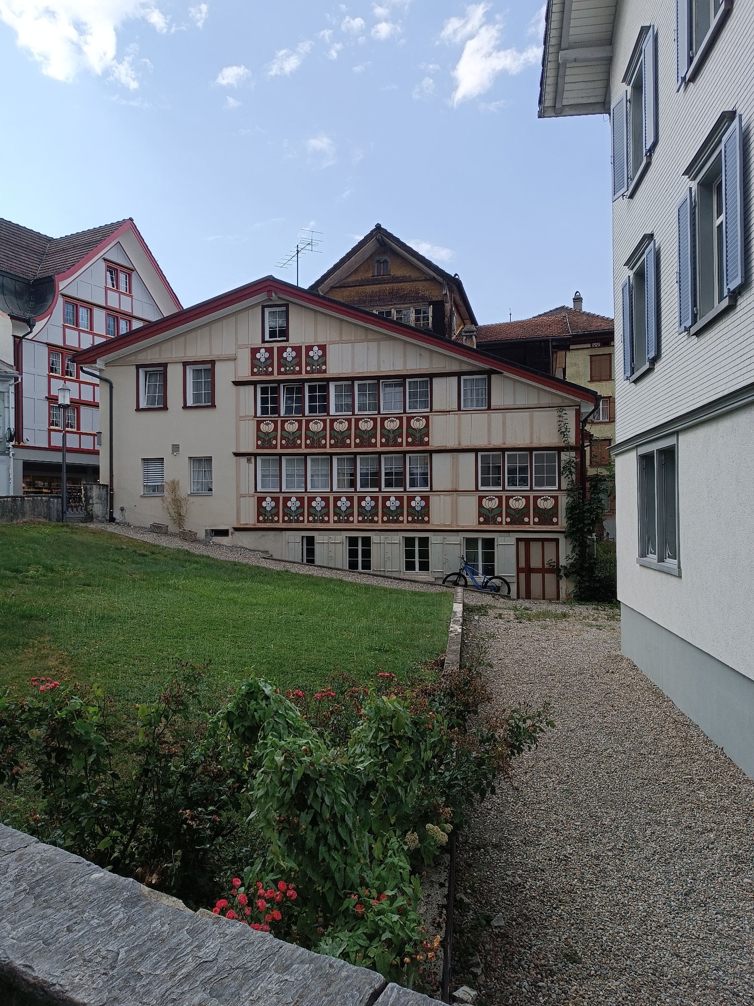 The image shows a street scene in Appenzell, Switzerland. In the foreground, a stone wall runs along the bottom of the frame, with a lush green lawn and flowering bushes on the other side. A gravel path leads up to a row of traditional Swiss houses. The houses are brightly painted, with decorative motifs of flowers and geometric patterns adorning their facades. Several windows are visible, some with closed shutters. A blue bicycle is parked near one of the houses. In the background, more houses and rooftops are visible, with a clear blue sky overhead. The overall impression is one of a charming and well-maintained historic village.