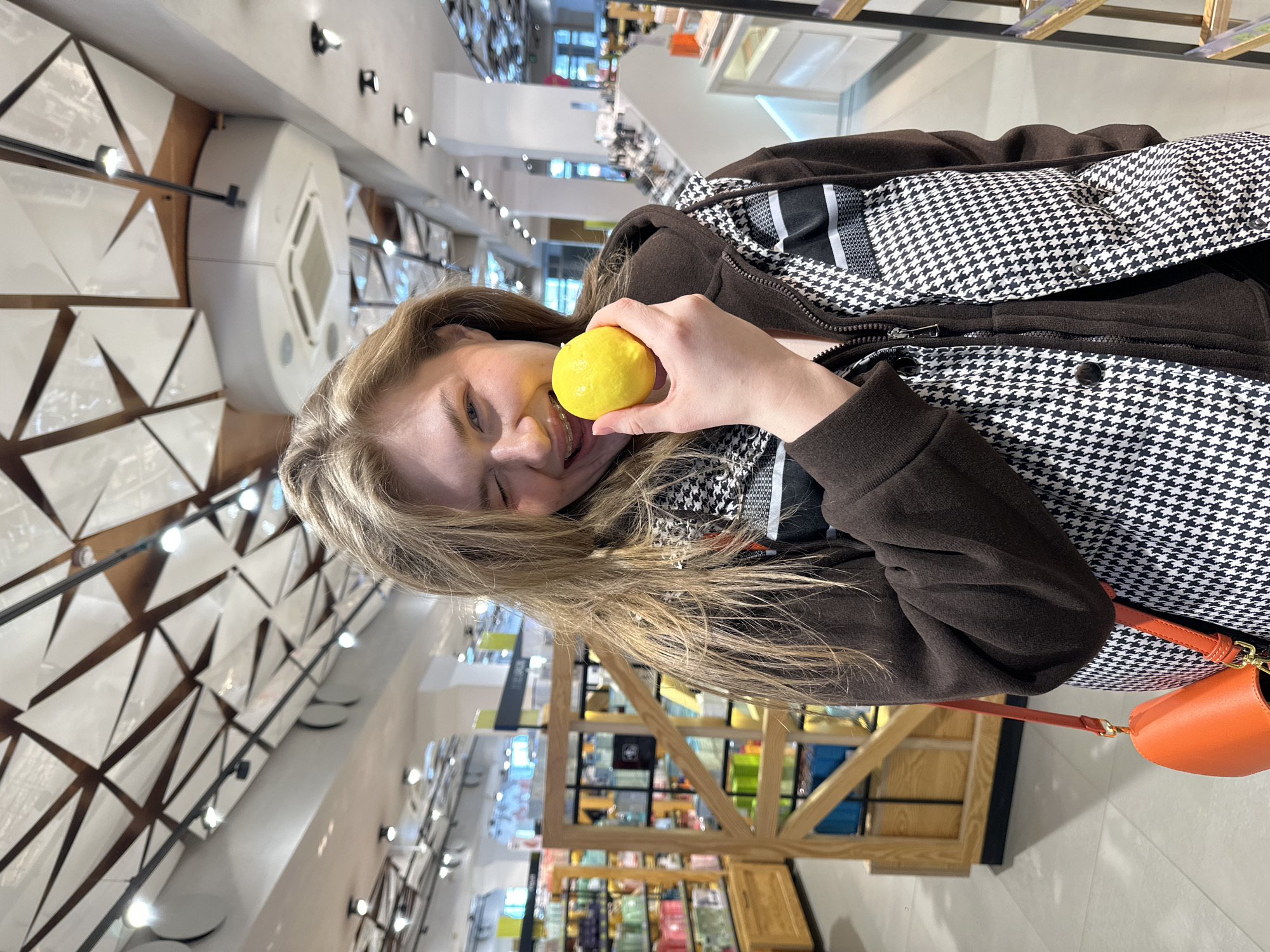 A young fair-skinned female with long blonde hair stands centrally in a retail store in Kazan, Russia. She is facing the camera, winking with her left eye, and smiling, revealing upper dental braces. Her right hand holds a bright yellow lemon close to her slightly open mouth, with yellow residue visible on her upper lip and right cheek. She wears a dark brown hooded jacket under a black and white houndstooth pattern vest. An orange shoulder bag with gold hardware hangs from her left shoulder. The background features a brightly lit store interior with a distinctive geometric ceiling composed of white triangular panels integrated with dark structural beams and track lighting. An HVAC unit is visible overhead. To the left, wooden display shelves are stocked with various colorful packaged goods. To the right, minimalist display counters are present. The specific ceiling design and modern retail environment strongly suggest a Bahetle supermarket location in Kazan, such as the one at Dekabristov Street, 131В.