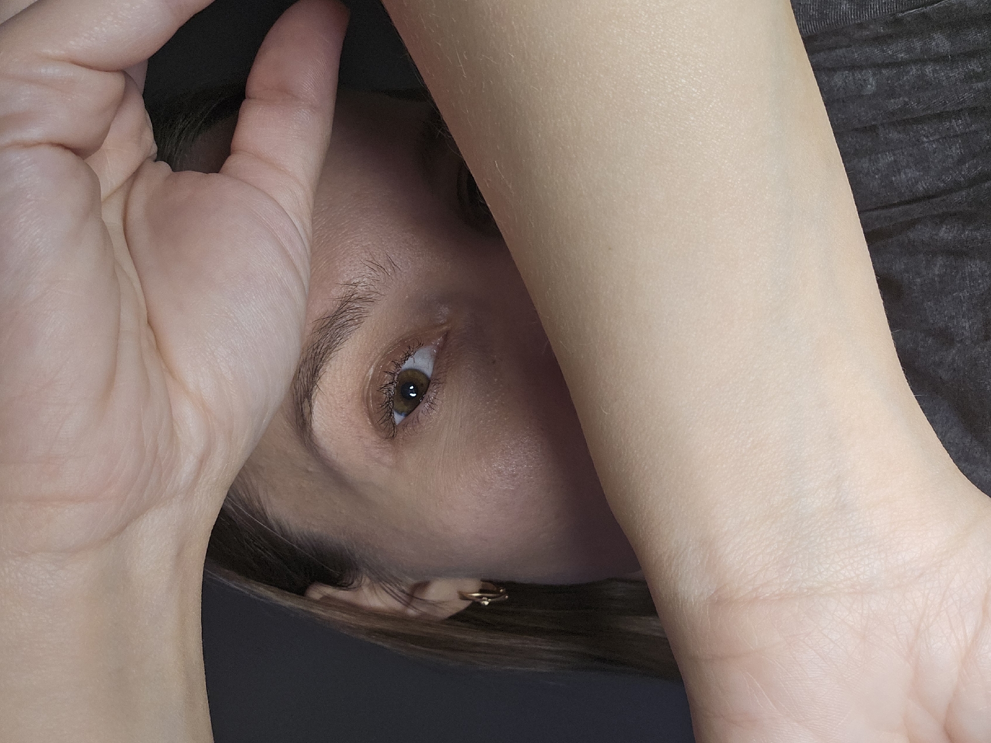 A close-up, rotated image shows a person's face partially obscured by their hands. One eye is visible, with an amber iris and visible eyelashes. Part of an earring is also visible. The person appears to be wearing a dark-colored garment. Skin tone is fair. The background is dark, obscuring details of the environment. There are no indicators within the image to suggest a specific location, including Chaikovsky, Russia.