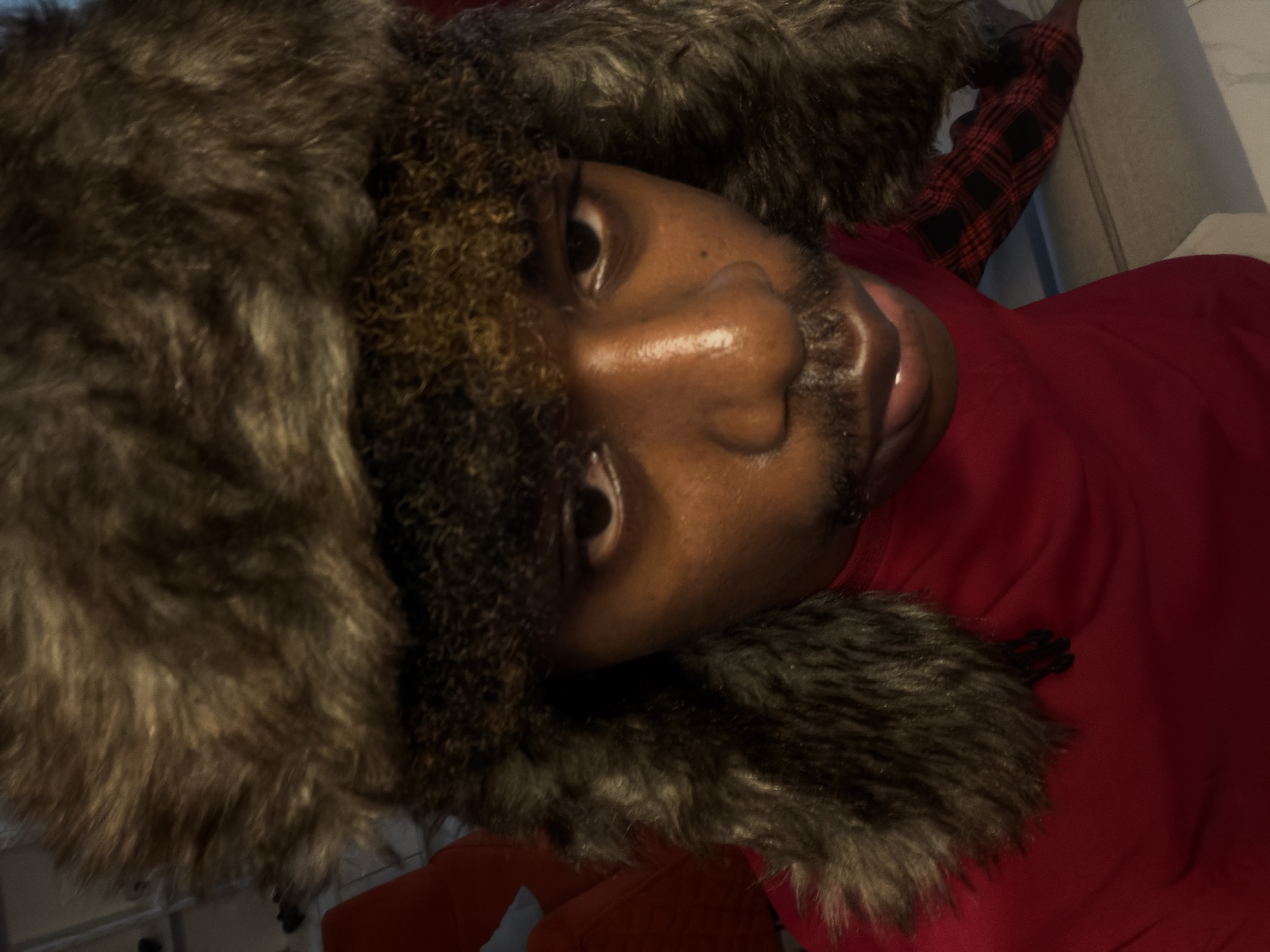 A medium shot, taken from above, shows a young Black person wearing a red long-sleeved shirt and a dark brown fur hat. The hat is a Ushanka-style hat with ear flaps and a fuzzy texture. The person has dark eyes, a mustache, and a faint goatee. The person's mouth is slightly open. The background is out of focus and appears to be indoors, with hints of red upholstery and a floor that looks like light gray tiles or concrete. The person's skin is smooth and appears slightly oily, reflecting the light. The overall lighting is dim. There is no indication of the specific location within Igboefon, Nigeria, only that the scene is presumed to be there.