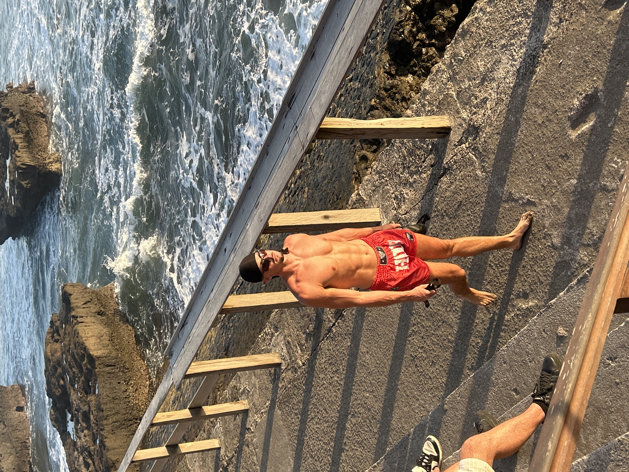 In North Kuta, Indonesia, a shirtless man wearing red athletic shorts and a black cap walks along a concrete pathway next to a wooden railing. The pathway is bordered by rocky terrain and the ocean. The man is looking to his right, and he is holding a dark object in his right hand. Another person's legs are visible on the left side of the image, wearing white shorts and black sneakers. The sun is casting long shadows across the pathway, indicating it is either early morning or late afternoon. The ocean is rough with white waves crashing against dark rock formations.