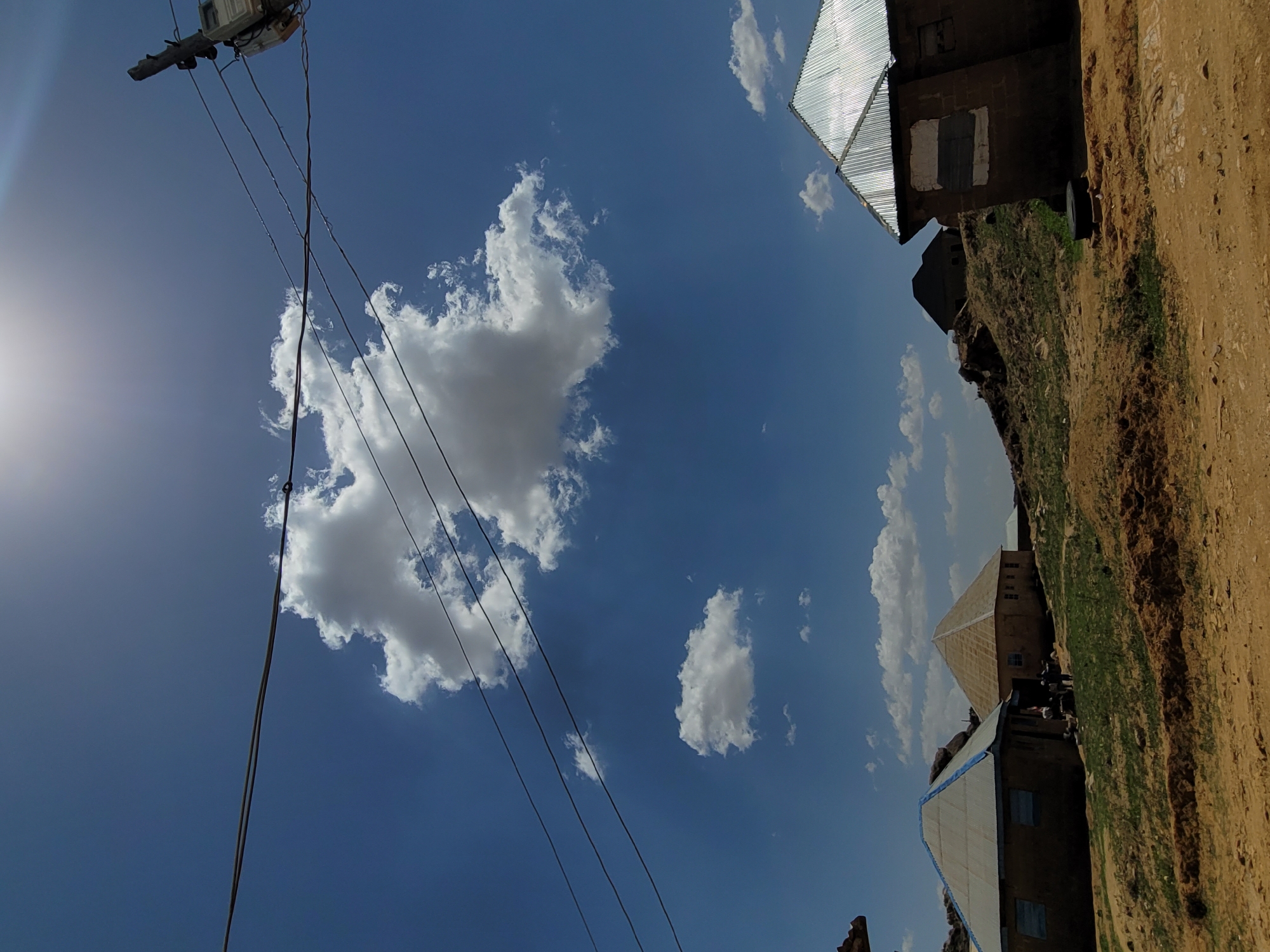 The image shows a rural landscape in Barakin Akawo, Nigeria, under a bright blue sky with scattered clouds. Electrical wires cross the frame. Buildings with corrugated metal roofs are seen, one appearing to be built on a rocky slope. The ground is a mix of dirt and sparse vegetation. No people are clearly visible.