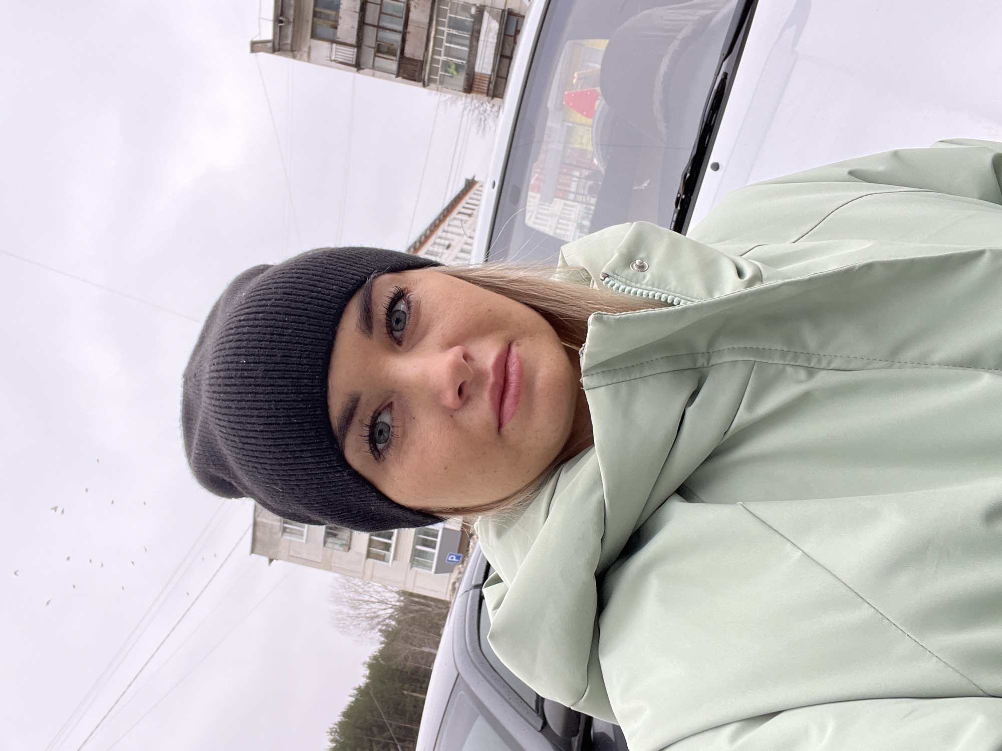 The image shows a young woman taking a selfie. She is wearing a black knit hat and a light green puffer jacket. She has fair skin and blue eyes. She is positioned in front of a white car, likely her own, as the image is taken from inside the vehicle looking out. The background reveals a typical urban scene, including multi-story apartment buildings common in Russian cities. Based on architectural styles and the presence of signage, the photo likely originates from Chaikovsky, Russia. The weather appears overcast and gray, as evidenced by the sky. Bare trees suggest a season of late autumn or early spring. A flock of birds can be seen in the background. A parking sign can be seen by the apartment building.