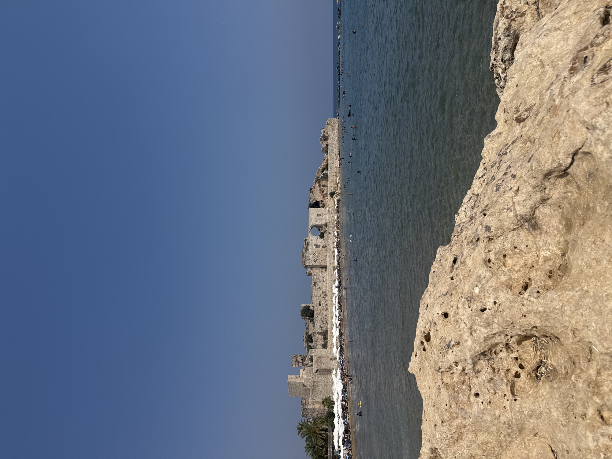 A clear, sunny day is depicted at a crowded beach in Erdemli, Turkey, with an ancient stone castle complex situated on the coastline. The foreground features a light-colored, weathered rock formation with numerous holes, partially obscuring the view. Beyond the rock, the sea is calm and a deep blue, with several people swimming and wading. The beach is lined with rows of white beach umbrellas and lounge chairs, with a few palm trees visible among them. The castle, constructed from large stone blocks, stretches along the coast and appears to be in a state of partial ruin, with some sections still intact, including arched openings and towers. A clear, cloudless blue sky dominates the upper portion of the image.