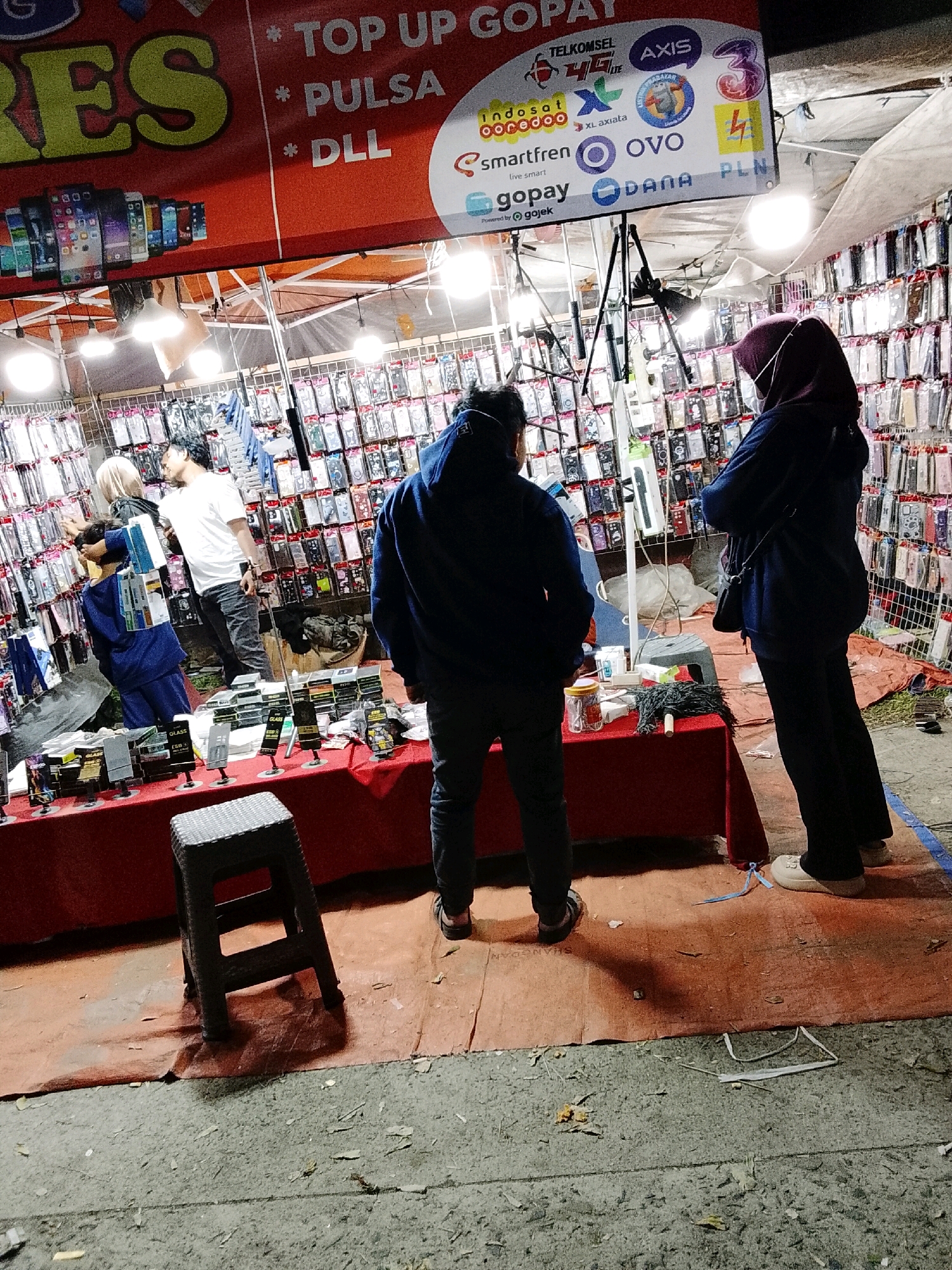 The image captures an outdoor market stall at night, likely in the city of Padang Dalom, Indonesia, given the presence of Indonesian brands and script on signs. A stall with a red tablecloth displays numerous phone cases and accessories. Above the stall, a large banner with yellow and red lettering proclaims "RESI" and "TOP UP GOPAY," along with smaller text mentioning "PULSA" (phone credit) and "DLL" (and so on). Several logos for telecommunication providers and payment services, such as Telkomsel, Axis, 3, XL Axiata, Smartfren, Ovo, Gopay, and Dana, are visible on signs.
Three individuals are present. Two are standing behind the stall, facing away from the camera, appearing to be customers or vendors. One is wearing a dark blue hooded sweatshirt and dark pants, while the other wears a white t-shirt and dark pants. Another person, dressed in a blue uniform, is partially visible on the left side of the stall, appearing to be interacting with the display. A woman wearing a dark hijab and dark pants is standing to the right of the stall, also facing away from the camera.
The ground is covered with an orange tarp, upon which a dark gray plastic stool rests. The background is filled with rows of phone cases, displayed in clear plastic packaging, suggesting a focus on mobile phone accessories. The lighting appears to be from overhead lamps, creating shadows on the ground. The overall scene depicts a commercial transaction or browsing activity at a night market.