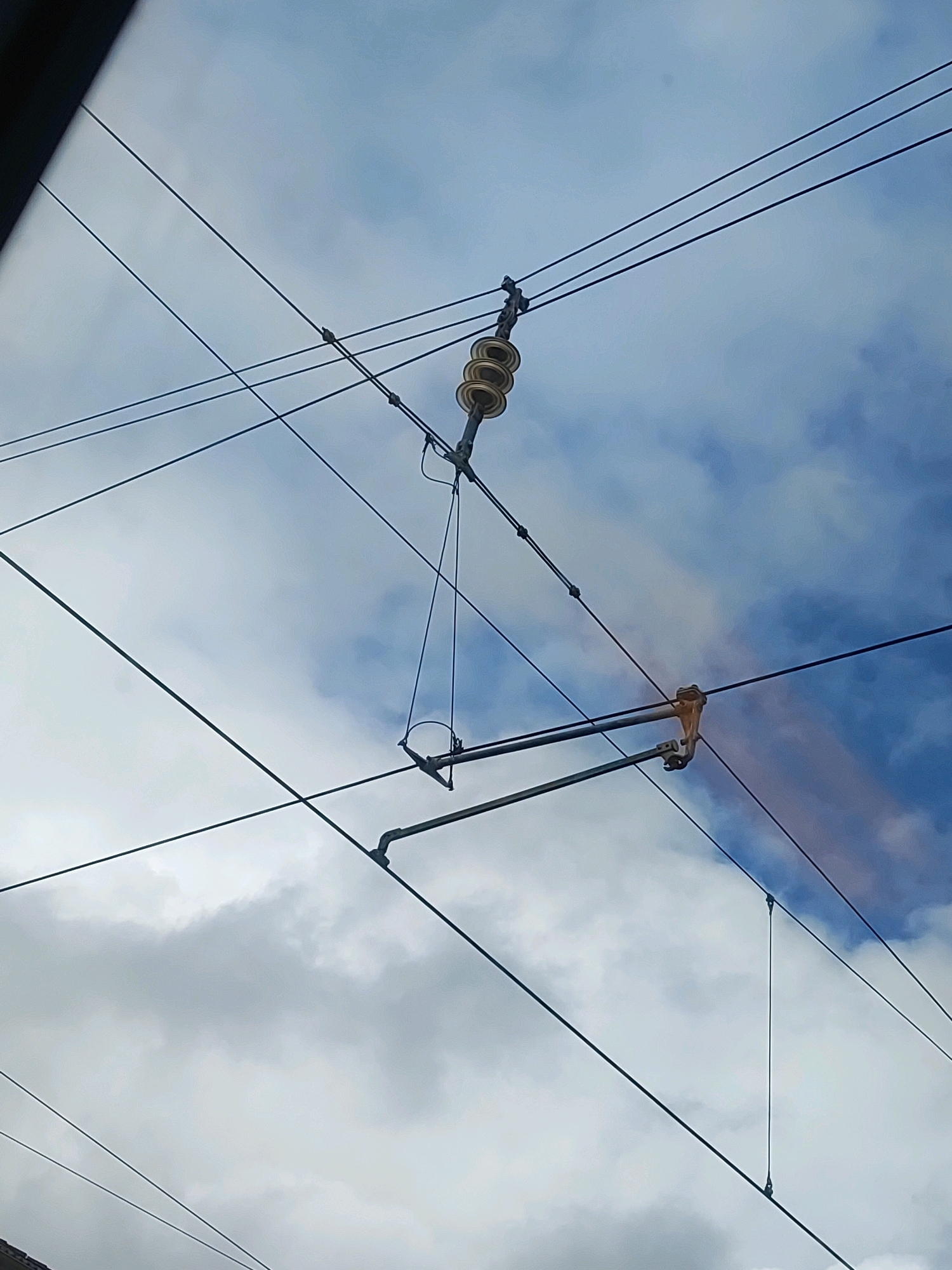 The image shows an overhead catenary system against a sky with clouds. Several dark wires are visible, some appearing to be tensioned and supported by insulators. One of these insulators, a beige, multi-ringed object, is prominently positioned on the left side of the frame. Other wires are suspended from a complex arrangement of metal poles and arms, with some wires crisscrossing each other. In the upper right, a section of a wire appears to have a reddish-brown discoloration. The sky is a mix of blue and white clouds, with the lighting suggesting it is daytime. This infrastructure is part of the railway line in Roßtal, Germany, likely used for powering electric trains.