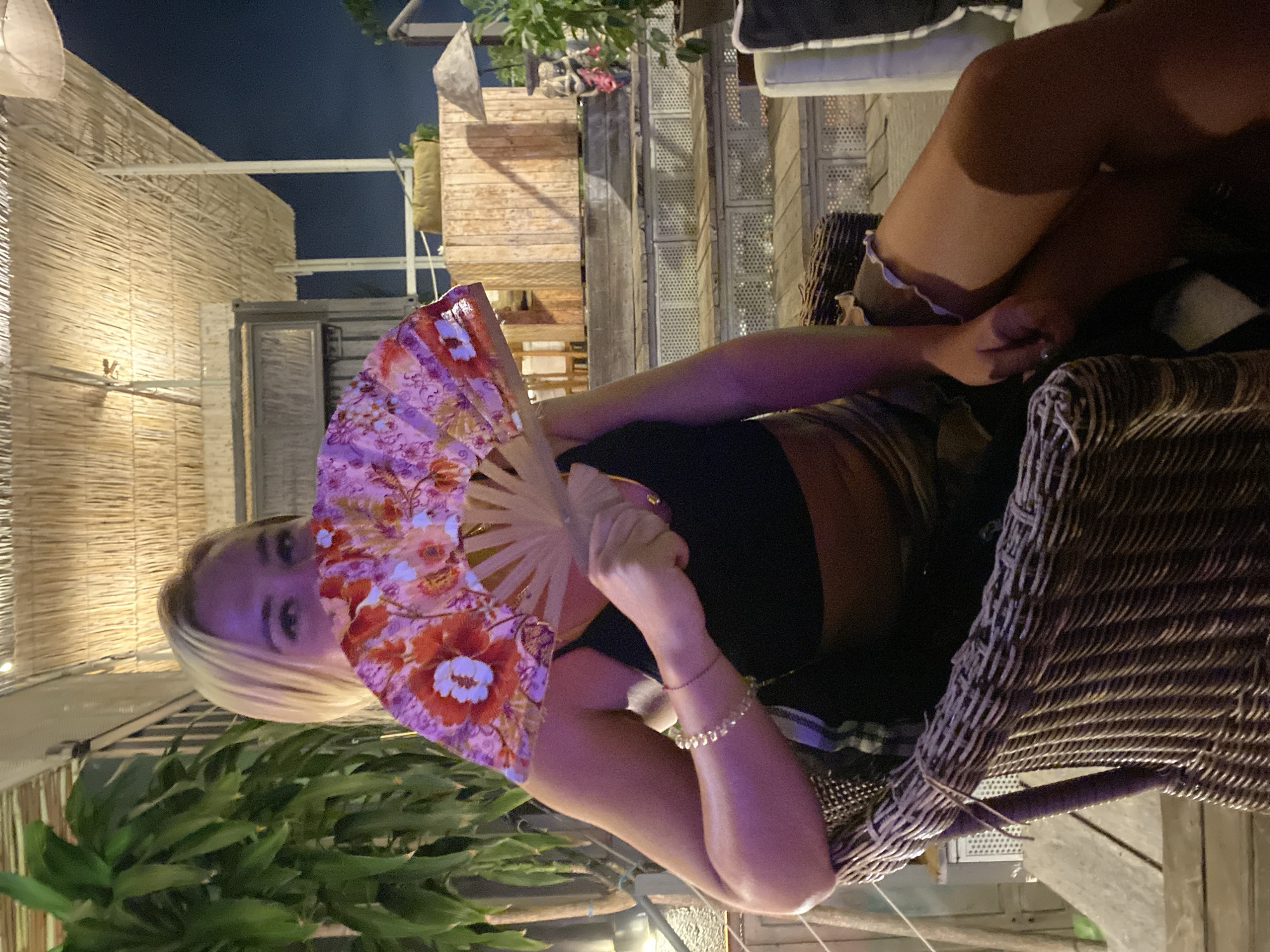 A person holds a floral fan in Mũi Né, Vietnam. They are seated in a wicker chair outdoors at night, with various buildings and plants in the background. The person wears jewelry on their wrist and a dark top.