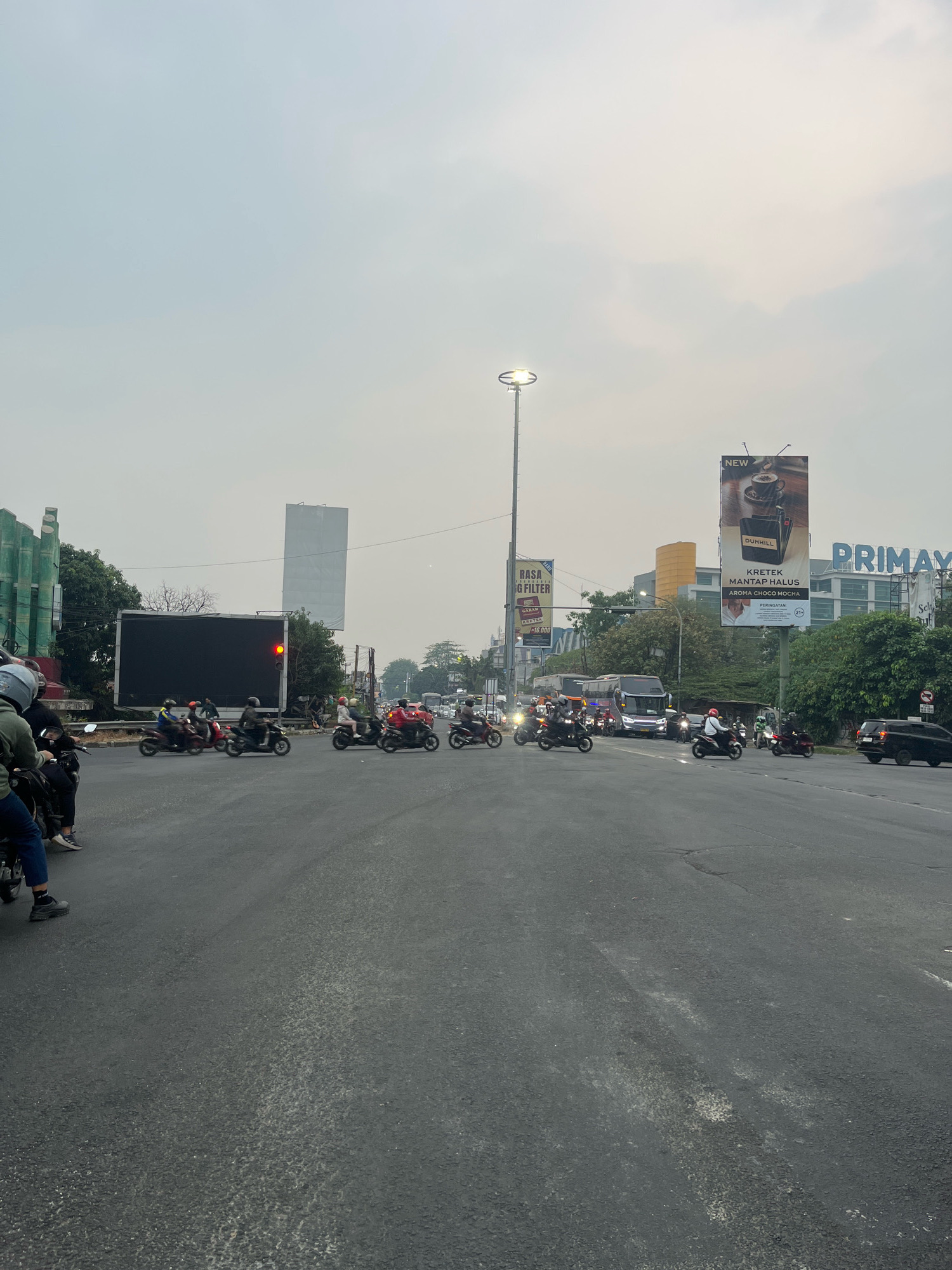 The image depicts a street intersection in Bekasi, Indonesia, under an overcast sky. A large, dark digital billboard is prominent on the left side of the road, displaying a single red traffic light. Several motorcycles are in motion, navigating the intersection. On the right side of the road, there are billboards advertising products, including one for "DUNHILL KRETEK MANTAP HALUS AROMA CHOCO MOCHA" and another for "PRIMAY". Buildings and trees line the sides of the road, and the overall impression is of a busy urban environment.