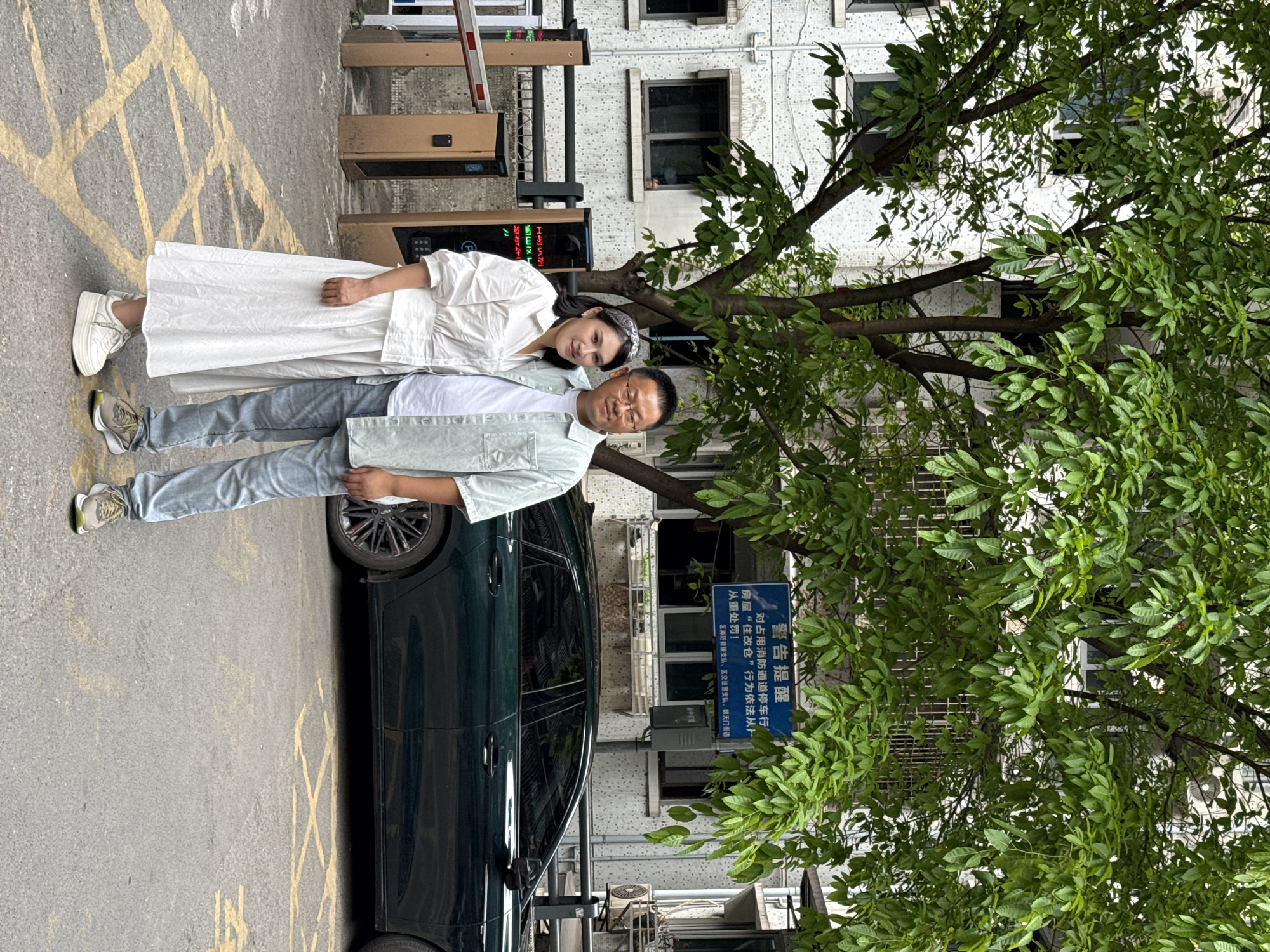Here is a description of the image:
A man and a woman are standing next to a dark green car on a street in Yuzhong District, China. The man is wearing glasses, a light green shirt, a white t-shirt, and blue jeans. The woman is wearing a white dress. A building is in the background. A sign with Chinese characters is visible near a tree.
