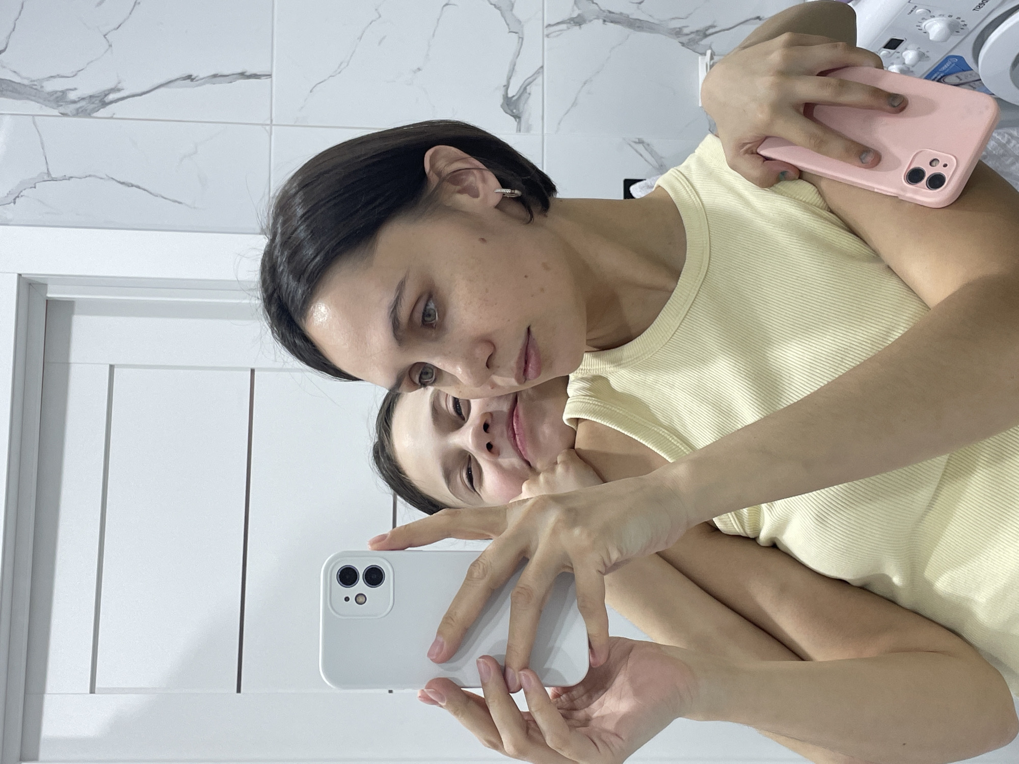 A woman with short dark hair is taking a selfie in a bathroom mirror. She is wearing a light yellow ribbed tank top. The reflection in the mirror shows another woman behind her, possibly the same woman, also smiling slightly. The woman in the foreground is holding a white smartphone, presumably to take the photo. Her other hand holds a pink smartphone. The background includes white marble-patterned tile on the wall, and a white door with vertical insets to the left of the woman. In the upper right corner of the frame, a white object with knobs is visible. The city of Izhevsk, Russia is not discernible in this image.