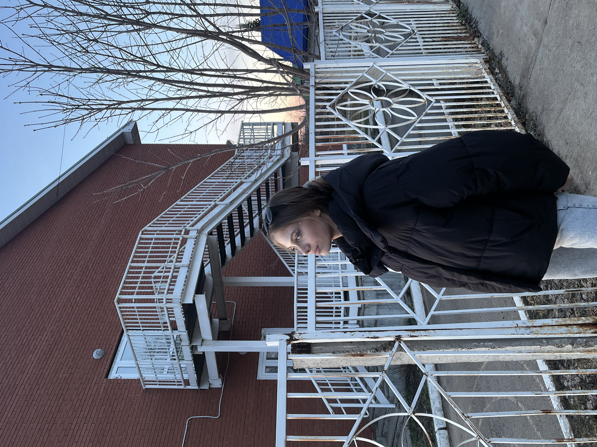 A woman wearing a black puffer jacket and light gray pants stands in front of a white metal fence adjacent to a brick building with a white metal fire escape. The building is located in Фоки, Russia, evidenced by geotagging information. A bare tree is visible behind the building and fence. The sky is pale blue and pink indicating it is dusk or dawn. The woman is looking down and slightly away from the camera. The metal fence shows some rust.