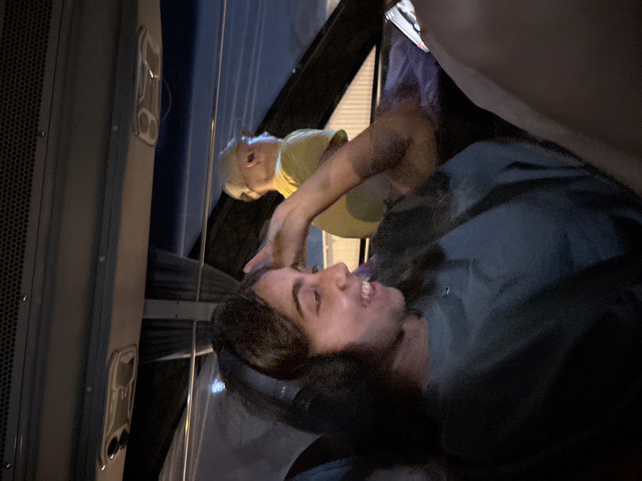A nighttime image depicts the interior of a vehicle, likely a bus or large van, in Samut Sakhon, Thailand.
In the foreground, a young adult female is seated in profile, facing right. She has dark, long hair and is wearing black over-ear headphones and a dark-colored short-sleeved top. Her facial expression shows a wide smile with visible teeth. Her right hand is raised with fingers splayed near her face. A mobile device with an illuminated screen is partially visible in the bottom right, held by her left hand.
In the background, an adult male is seated further back and to the right, viewed in profile facing left. He wears a light-colored baseball cap, eyeglasses, and an in-ear wireless earbud in his right ear. He is dressed in a light yellow short-sleeved shirt. His right arm rests on a purple object, possibly a blanket or bag, on the adjacent seat.
The vehicle's interior is dimly lit by artificial overhead sources, with visible ventilation panels and fixtures. Through the vehicle's window, partially obscured by a dark curtain, the exterior is dark, with faint white vertical structures and light sources indicating an outdoor nighttime environment. No specific identifying landmarks for Samut Sakhon are discernible in the image.