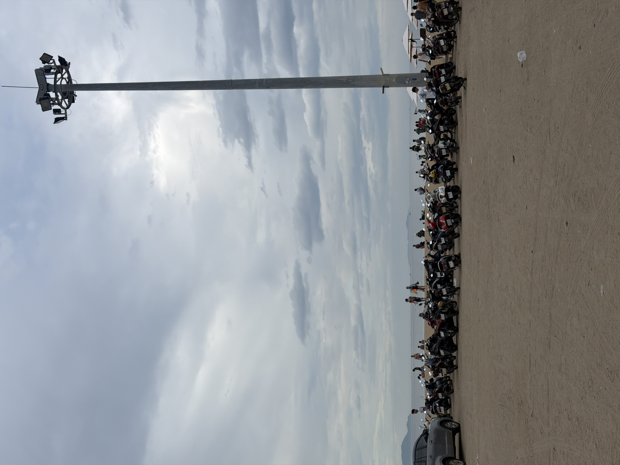 The image depicts a cloudy day at a beach parking lot in Nha Trang, Vietnam. A tall, metal light pole stands prominently on the right side of the frame. In the foreground, a sandy area is filled with a large number of parked motorcycles, varying in color from black to red and white. A silver car is visible on the far left. Beyond the parking lot, the beach is populated with several people, some standing and others appearing to be walking or relaxing. The sea stretches out in the background, with distant hills or landmasses visible on the horizon under the overcast sky. White beach umbrellas are set up further down the beach, suggesting a recreational area.