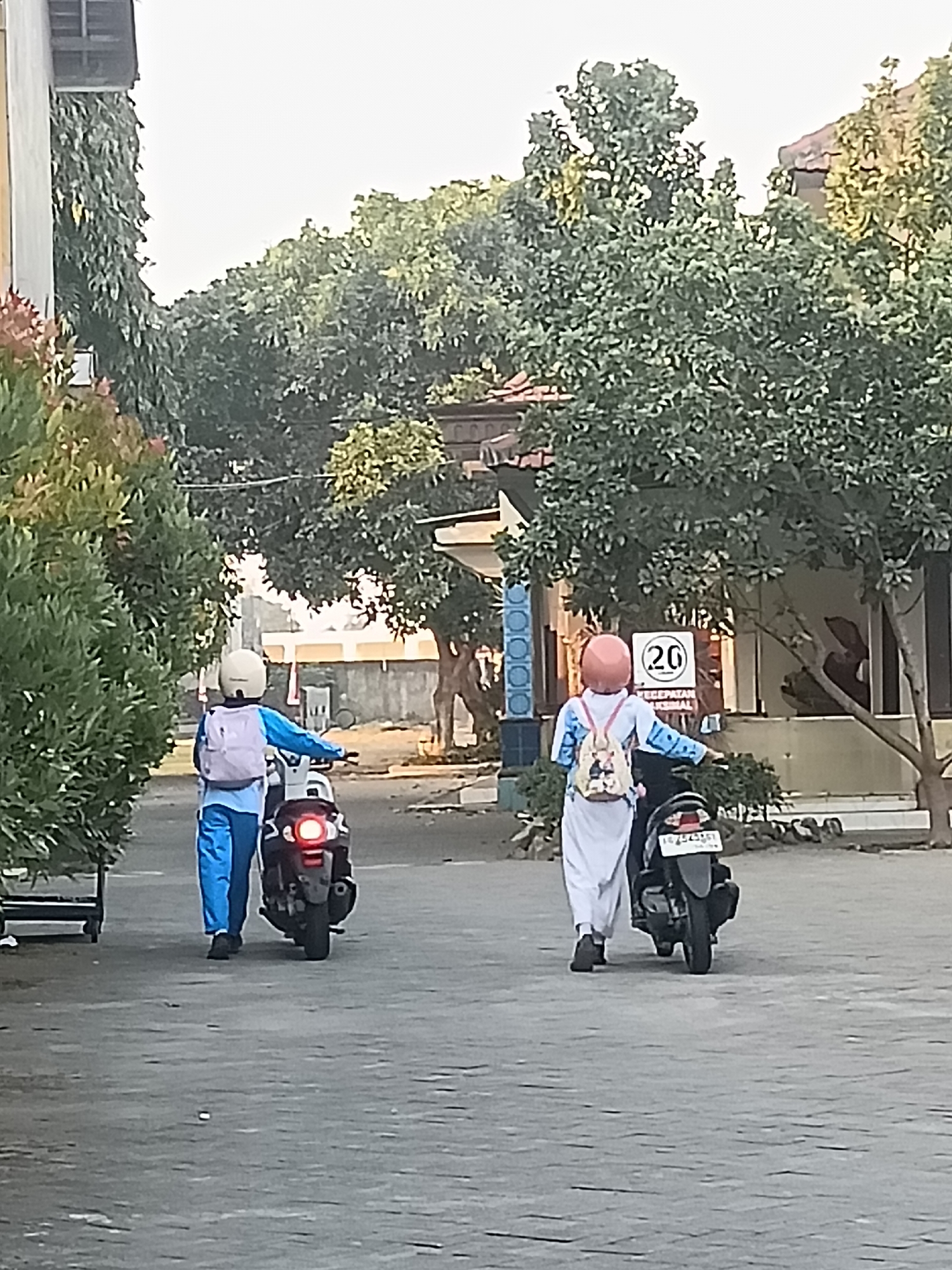 In Kediri City, Indonesia, two individuals, likely students, are walking their motorcycles on a paved path. The person on the left is wearing a light blue long-sleeved shirt and blue pants with a white helmet and a light-colored backpack. They are walking a dark-colored motorcycle with a red taillight. The person on the right is wearing a light-colored long-sleeved shirt with blue accents and light-colored pants, a pink helmet, and a patterned backpack. They are walking a dark-colored motorcycle with a license plate that reads "AG 5253 SJ". Both individuals are walking away from the viewer. The environment is an outdoor setting with lush green trees and some buildings in the background. A speed limit sign indicating "20" is visible on a post to the right of the image. The ground is a grey, textured pavement.