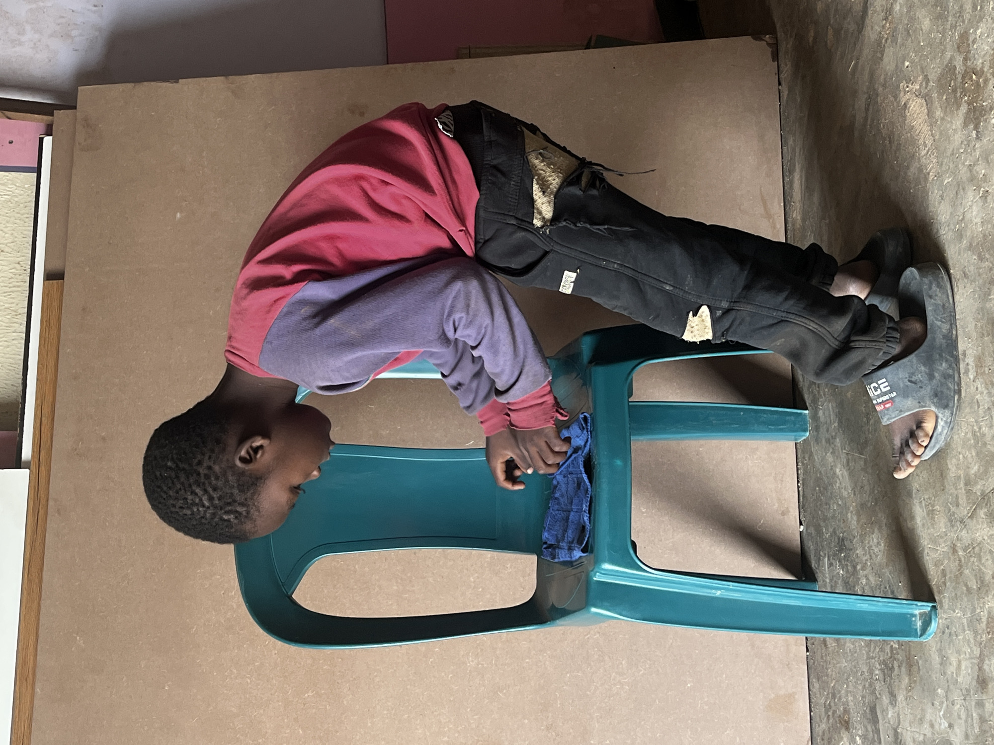 A young boy, with dark skin and short, dark hair, is shown in a standing, bent-over position in front of a teal plastic chair. He is wearing a long-sleeved shirt that is red on the sleeves and upper chest, transitioning to purple on the lower sleeves and torso. His pants are black, with distressed patches on the right thigh and knee, exposing a tan, textured material underneath. He is wearing dark gray slide sandals. In his hands, he holds a folded blue cloth and appears to be cleaning the seat of the chair. The chair is placed on a concrete floor with visible texture and some debris. Behind the boy and the chair is a large, light brown board leaning against a wall. To the right of the board, a portion of a wall is visible, exhibiting a textured, uneven surface. The background also shows stacked items, possibly boards or large sheets of material. The lighting suggests an indoor environment with ambient light. The scene depicts a domestic or communal space, likely within a residential area or workshop in Jos, Nigeria.