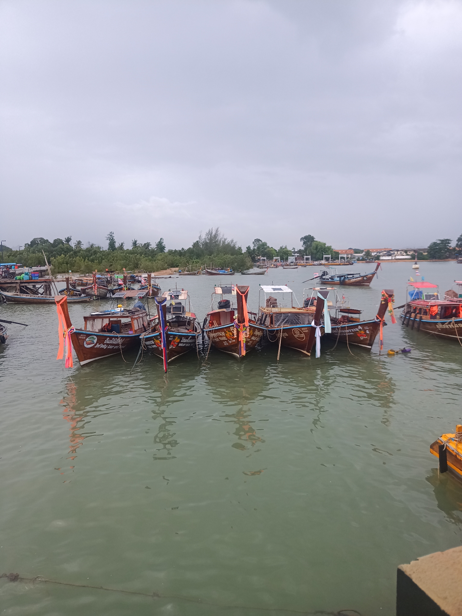 This is an outdoor scene in Ao Nam Mao, Thailand on an overcast day. The focus is on a group of wooden longtail boats docked or moored close together. The boats are decorated with colorful ribbons and flags at their prows. The water is calm with slight ripples, reflecting the boats and sky. In the background, there is a shoreline with green trees and buildings. The foreground includes a concrete structure, suggesting a pier or dock. Several other boats are in the water further from the main group, scattered across the bay. No people are visible in the immediate vicinity of the boats.