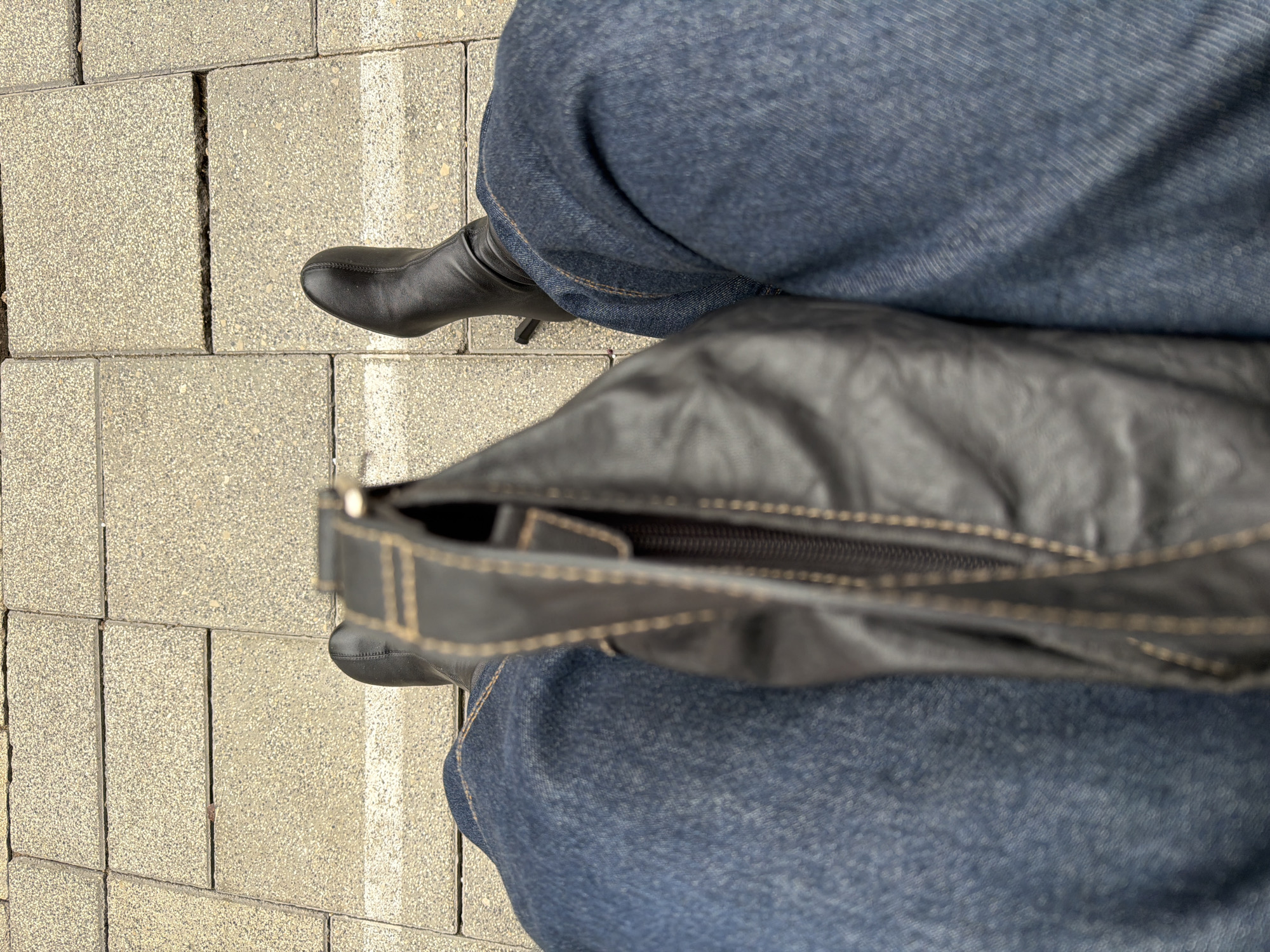 The image presents an overhead view of a person standing on a paved surface, likely a sidewalk or pedestrian area. The person is wearing dark blue jeans and black, high-heeled ankle boots. A black leather handbag is positioned in front of them, obscuring part of their lower body. The background shows light gray paving stones with narrow white lines between the rows. There is no definitive information in the image to confirm its location as Tel-Aviv, Israel. The image contains no discernable information about interactions.