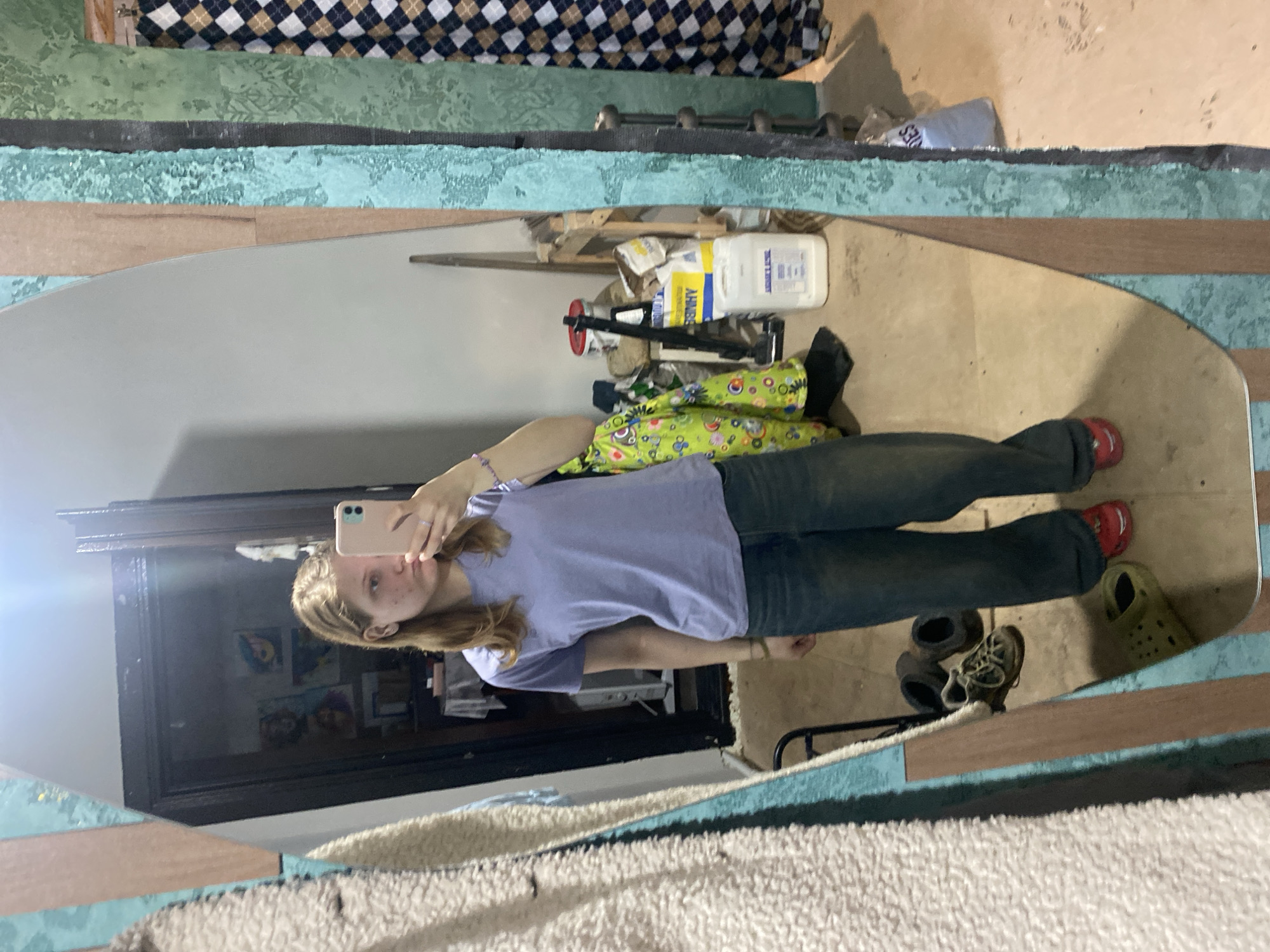 A reflection in a large oval mirror depicts a woman taking a selfie with a pink phone. She is wearing a light purple t-shirt and dark jeans with red shoes. The reflection reveals part of a room: the floor shows a pair of shoes, green clogs, and clutter. Behind her, visible through the mirror, is a window or door with framed pictures on the wall. A bed with a patterned cover and a textured surface are also visible within the mirror's reflection. The mirror itself is framed with alternating horizontal bands of blue and brown colors. The location is identified as the city of Chaikovsky, Russia.