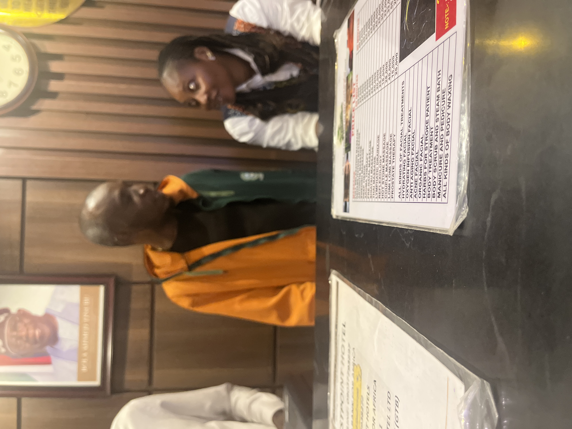 The image shows an interior scene at the "WESTPOINT HOTELS" in Abuja, Nigeria, likely a reception area. A portrait of a man, identified by text below as "BOLA AHMED TINUBU," hangs on a wood-paneled wall. In the foreground, a dark counter is visible with two price lists laid out. On the left, a partial price list for "WESTPOINT HOTELS" is visible, mentioning "BANKS FOR AFRICA." On the right, a more complete price list details various massage therapies and facial treatments with corresponding prices in what appears to be Nigerian Naira.
Two individuals are partially visible behind the counter. On the left, a person in a white top is mostly obscured. In the center, a man wearing an orange and green hooded jacket is facing towards the right, appearing to be in conversation with a woman. The woman, on the right, is wearing a white shirt and a patterned vest, and is looking towards the man. Her hair is dark and styled. The lighting is warm, suggesting indoor lighting. The overall environment appears to be a commercial establishment, possibly a hotel lobby or spa reception.